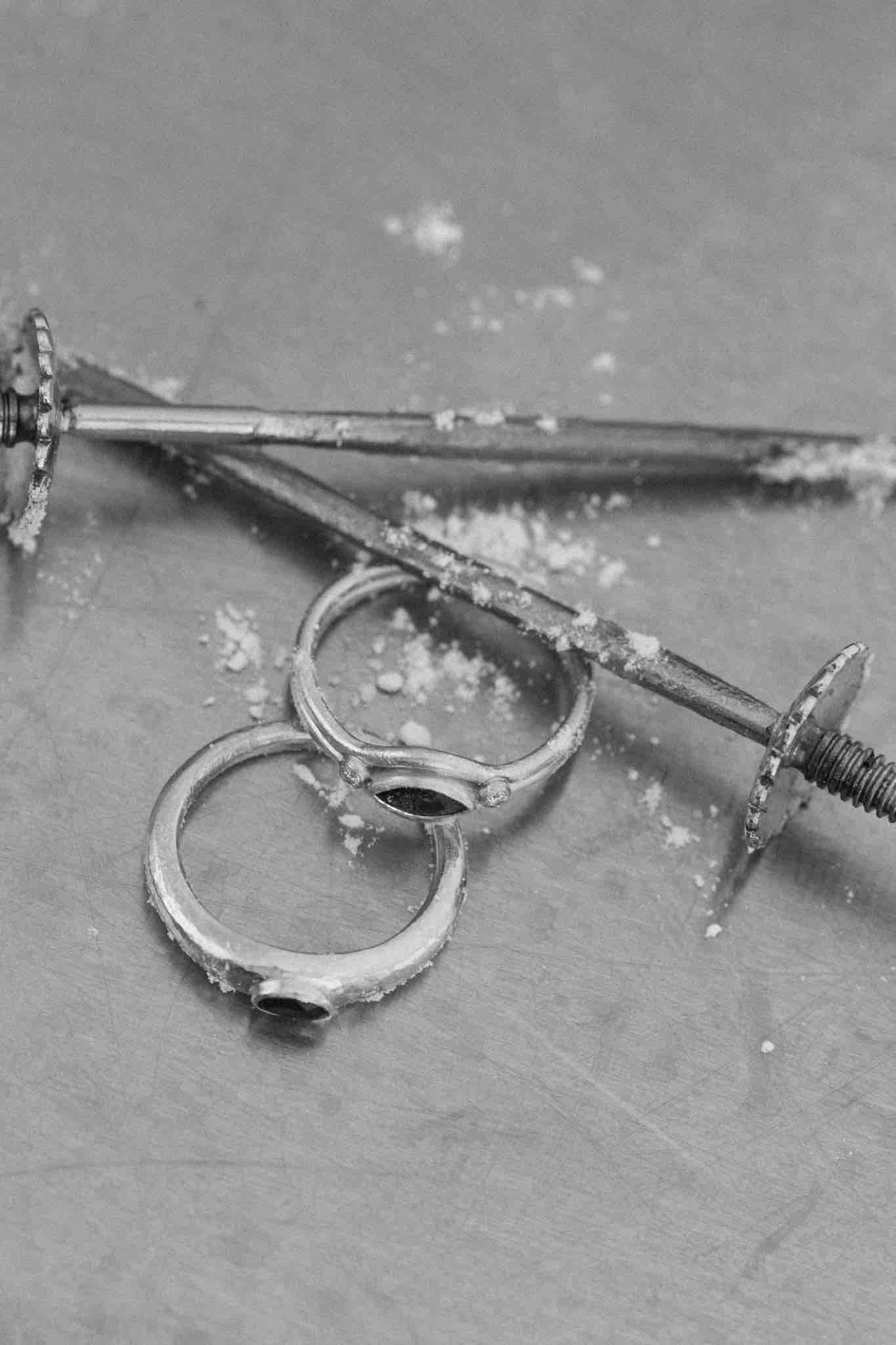 Two solid gold rings are laid next to silver sword skewers, dusted with icing powder on an industrial surface.