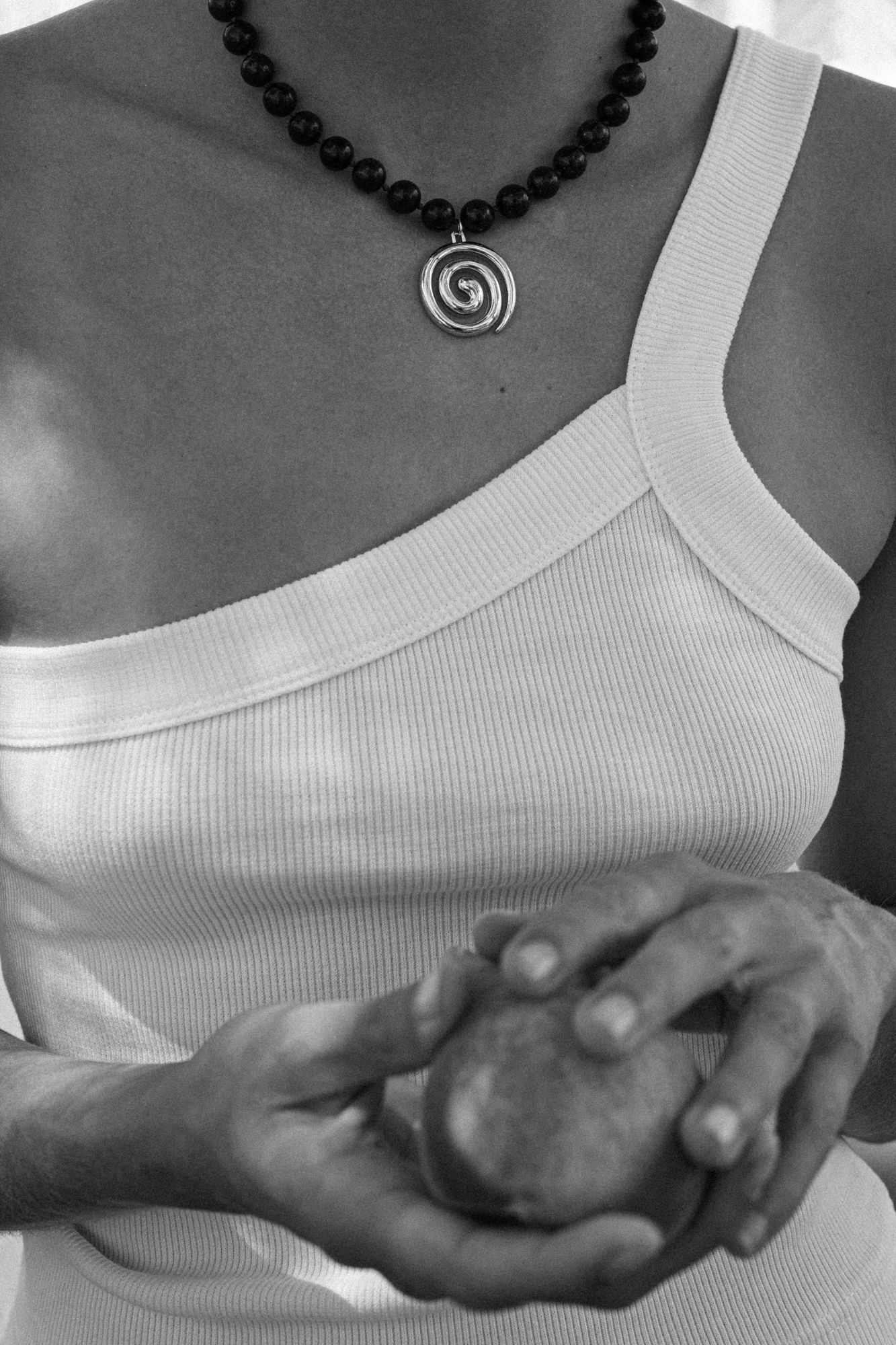 A black and white image of a model clasping a peach, while wearing a beaded necklace with a swirly sterling silver pendant.