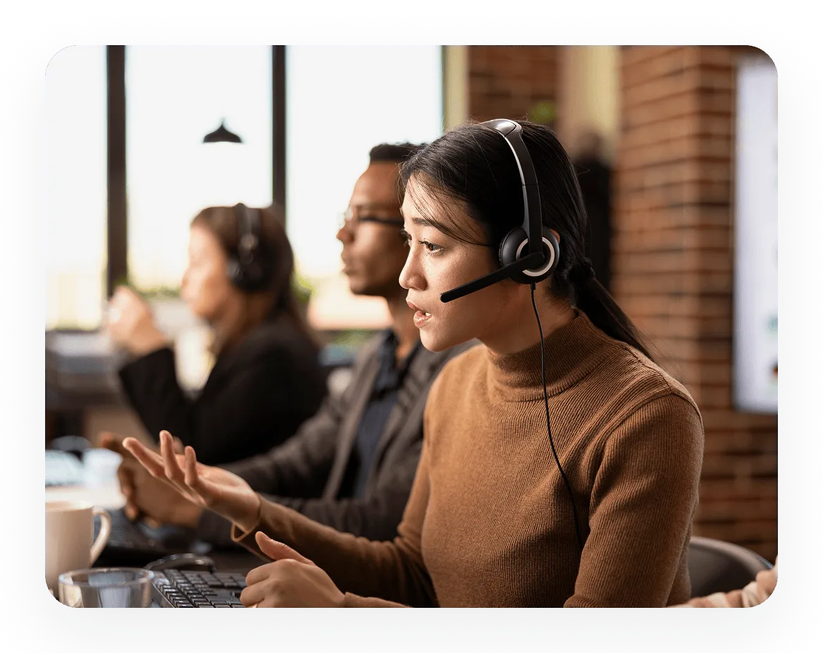 Woman with headset talking and gesturing with her hand in an office with colleagues working at computers in the background.