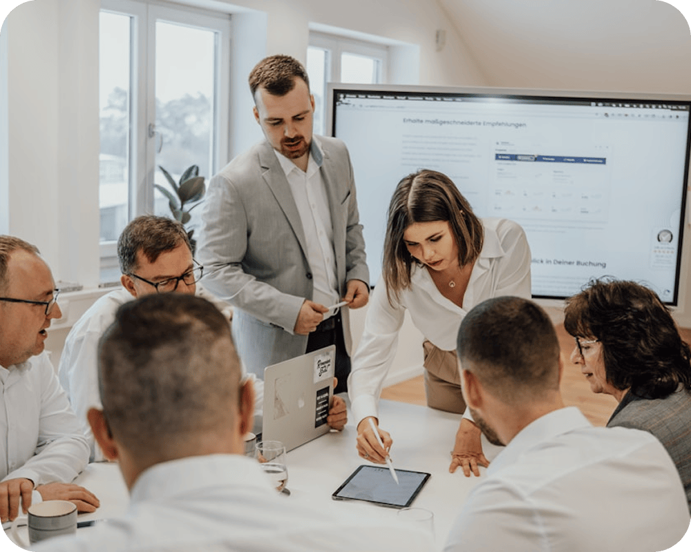 A group of six professionals having a meeting in an office, with a woman pointing at a tablet and a man standing nearby.