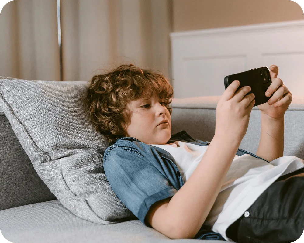 Young boy with curly hair lying on a gray couch playing on a smartphone.