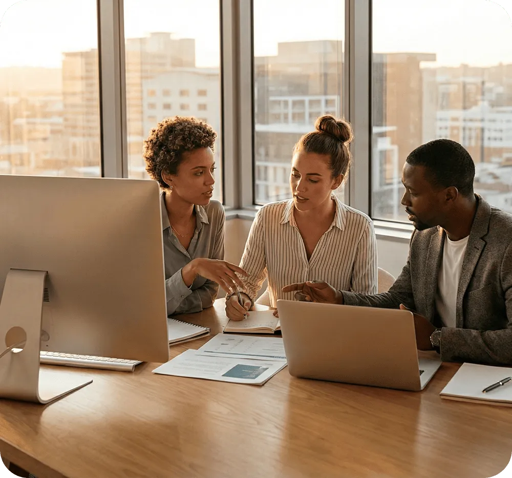 Three diverse colleagues discussing work around a desk with a computer, laptop, and documents in a modern office.