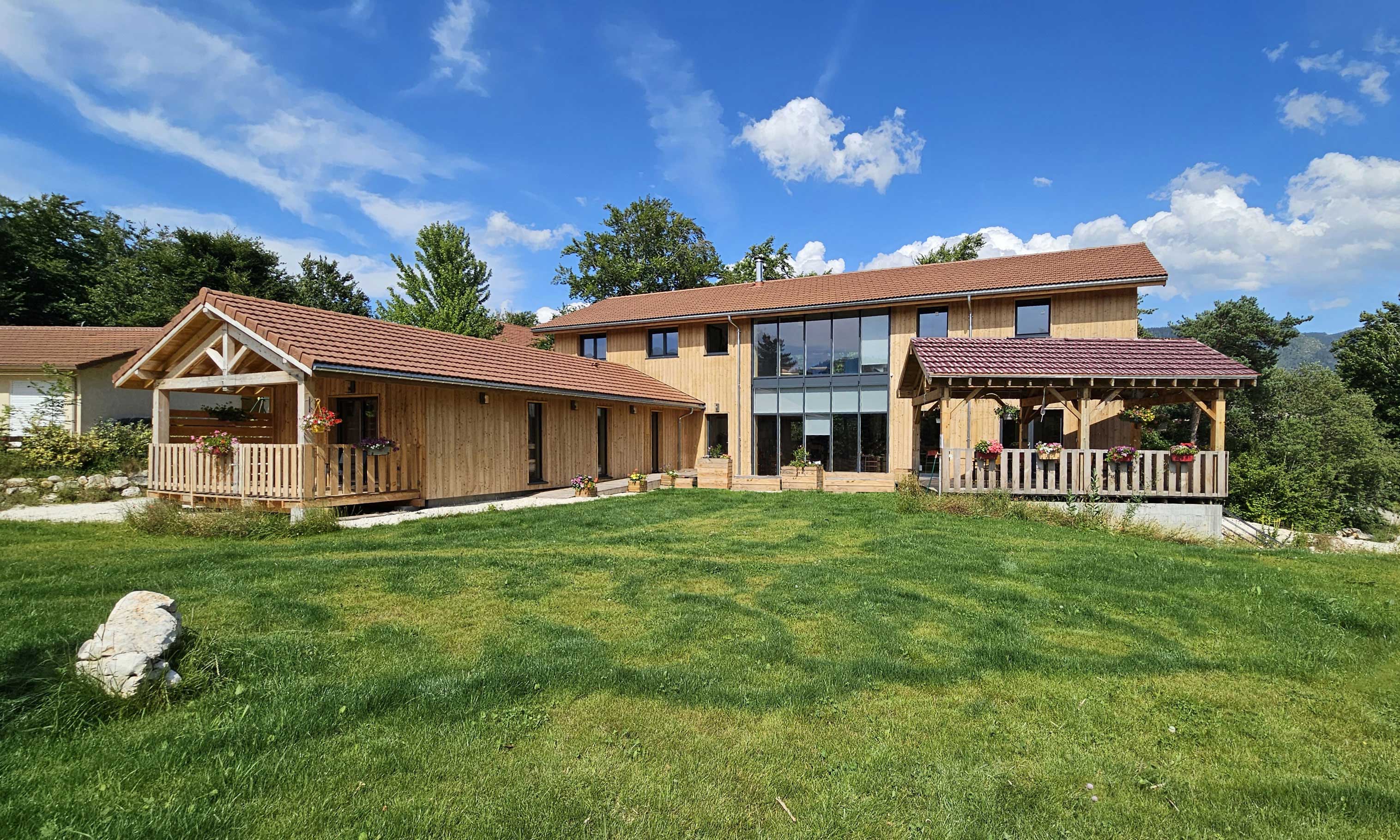 Grande maison en bois avec plusieurs toits en tuiles rouges, balcon fleuri et pelouse verte sous un ciel bleu avec quelques nuages.