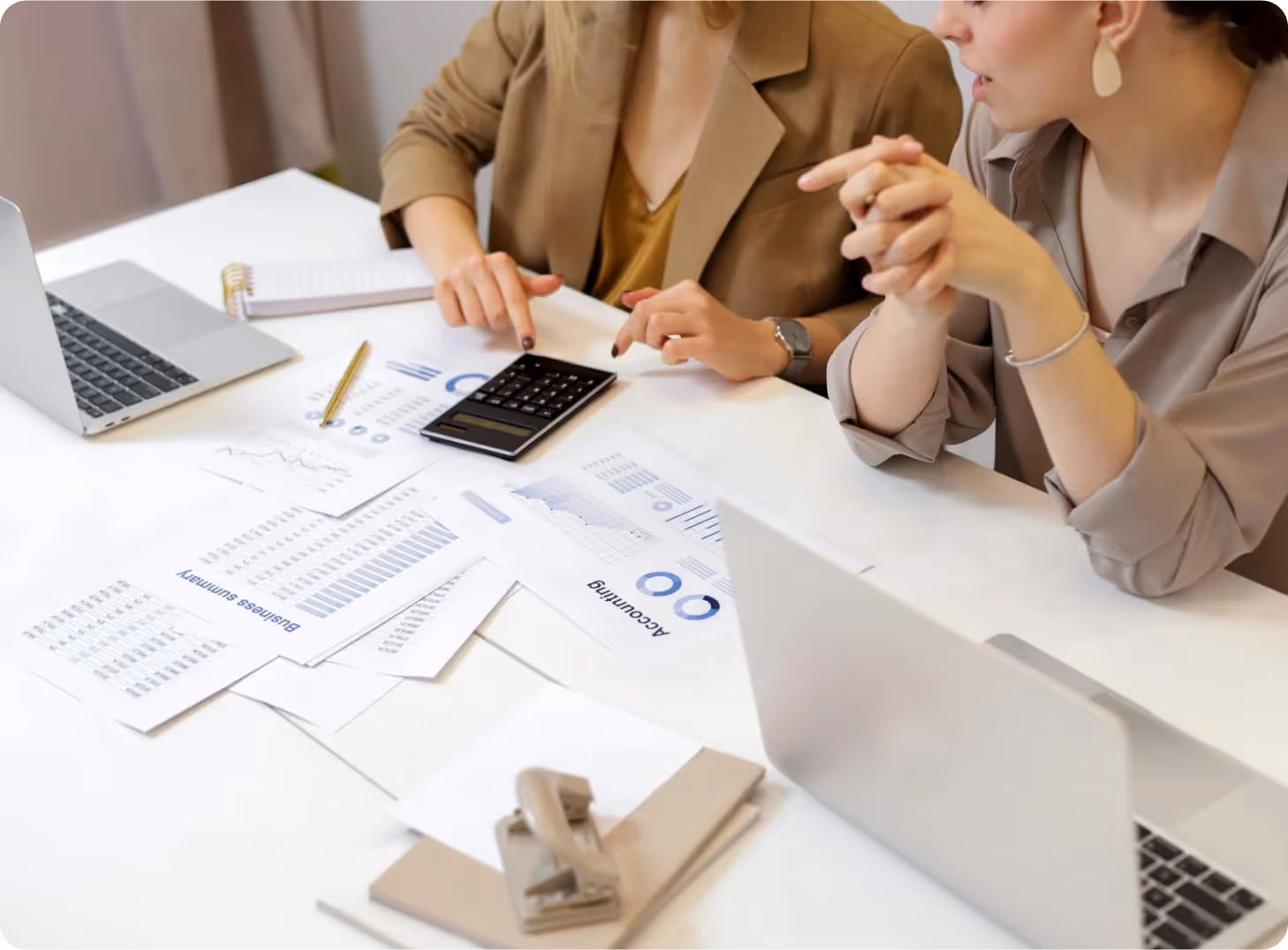 Two women reviewing financial documents and using a calculator at a white desk with laptops.