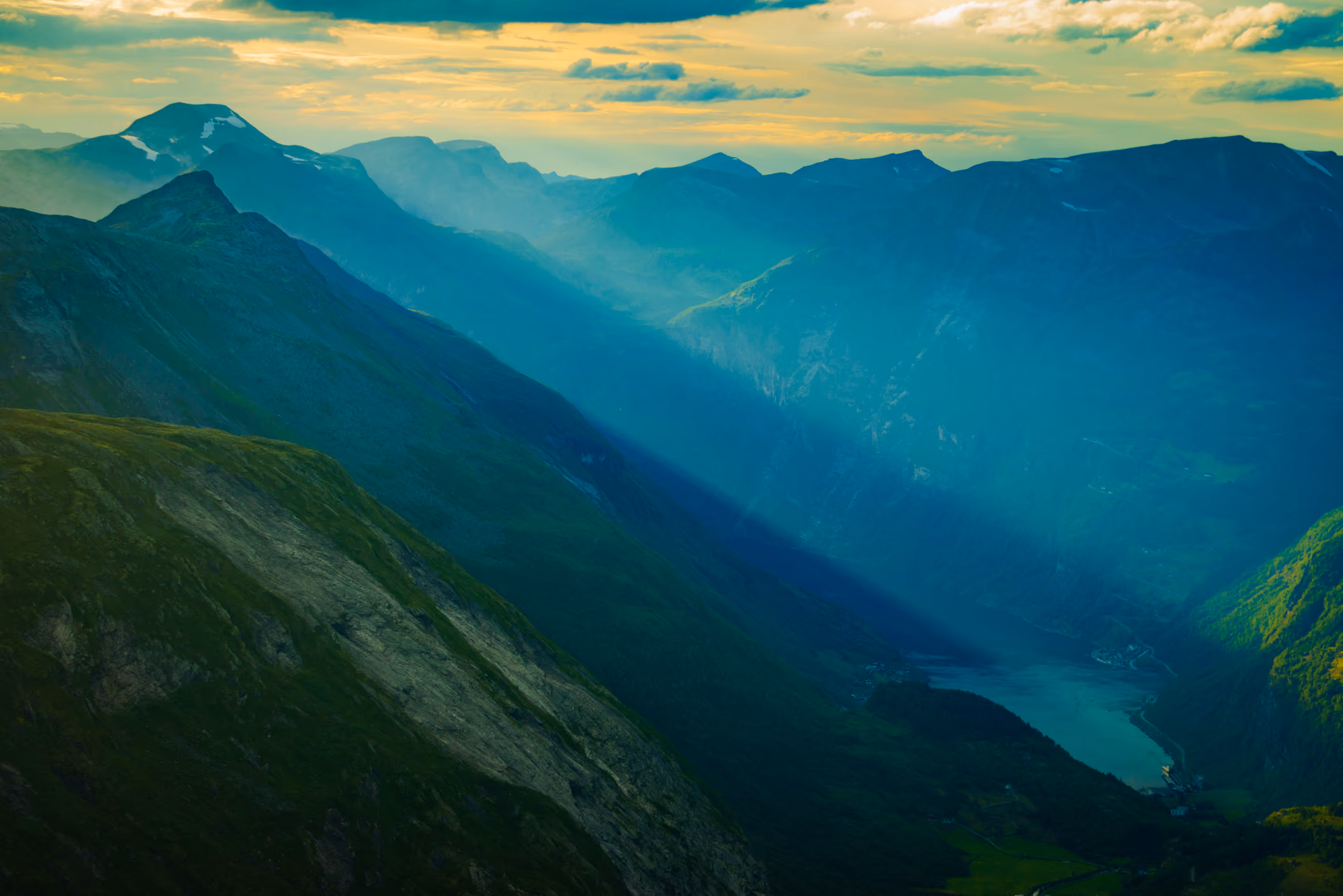Sunlight casting shadows over a green mountainous valley with a river or fjord running through it under a partly cloudy sky.