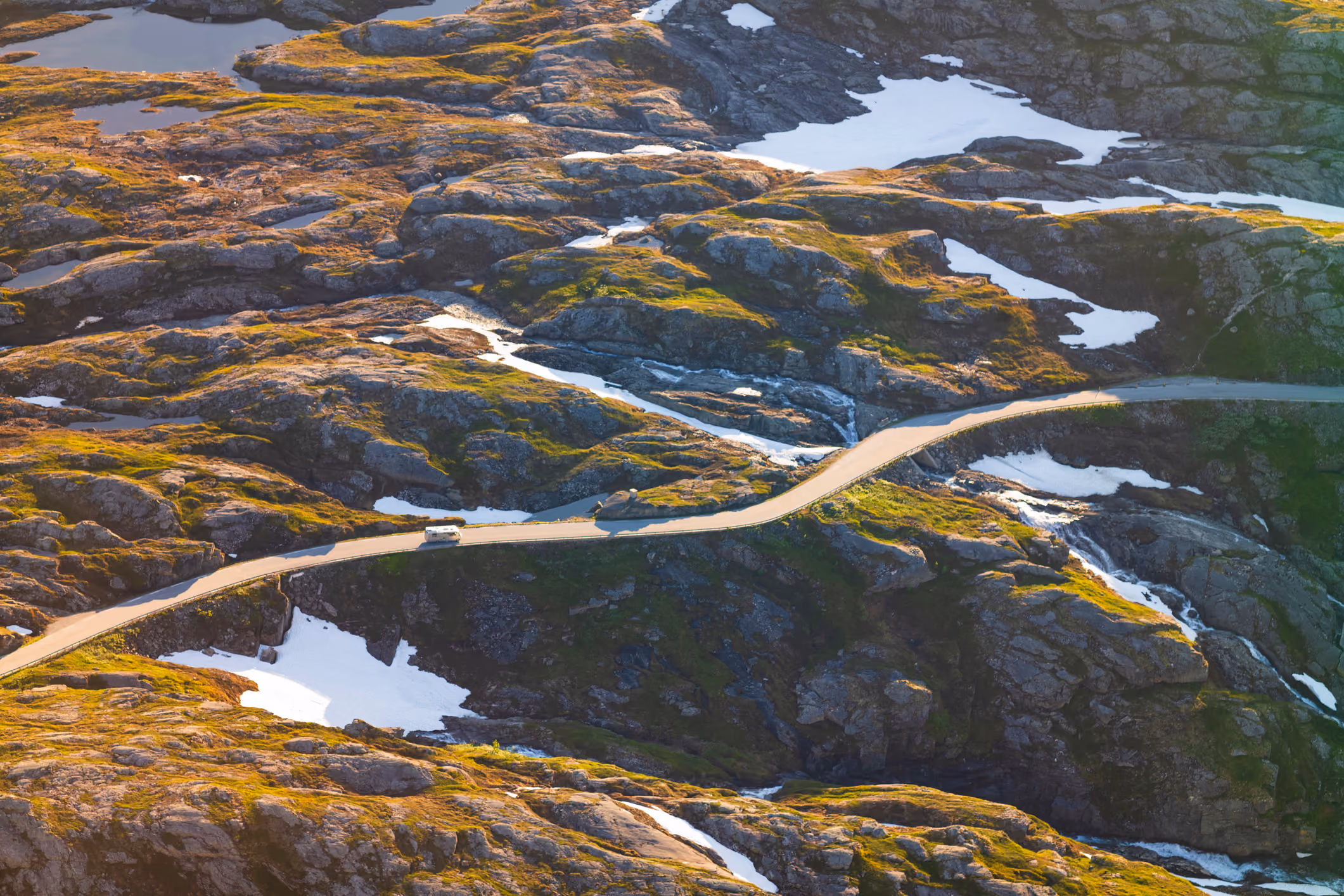 A winding road crosses rocky, snow-patched terrain with a white camper van driving on it.