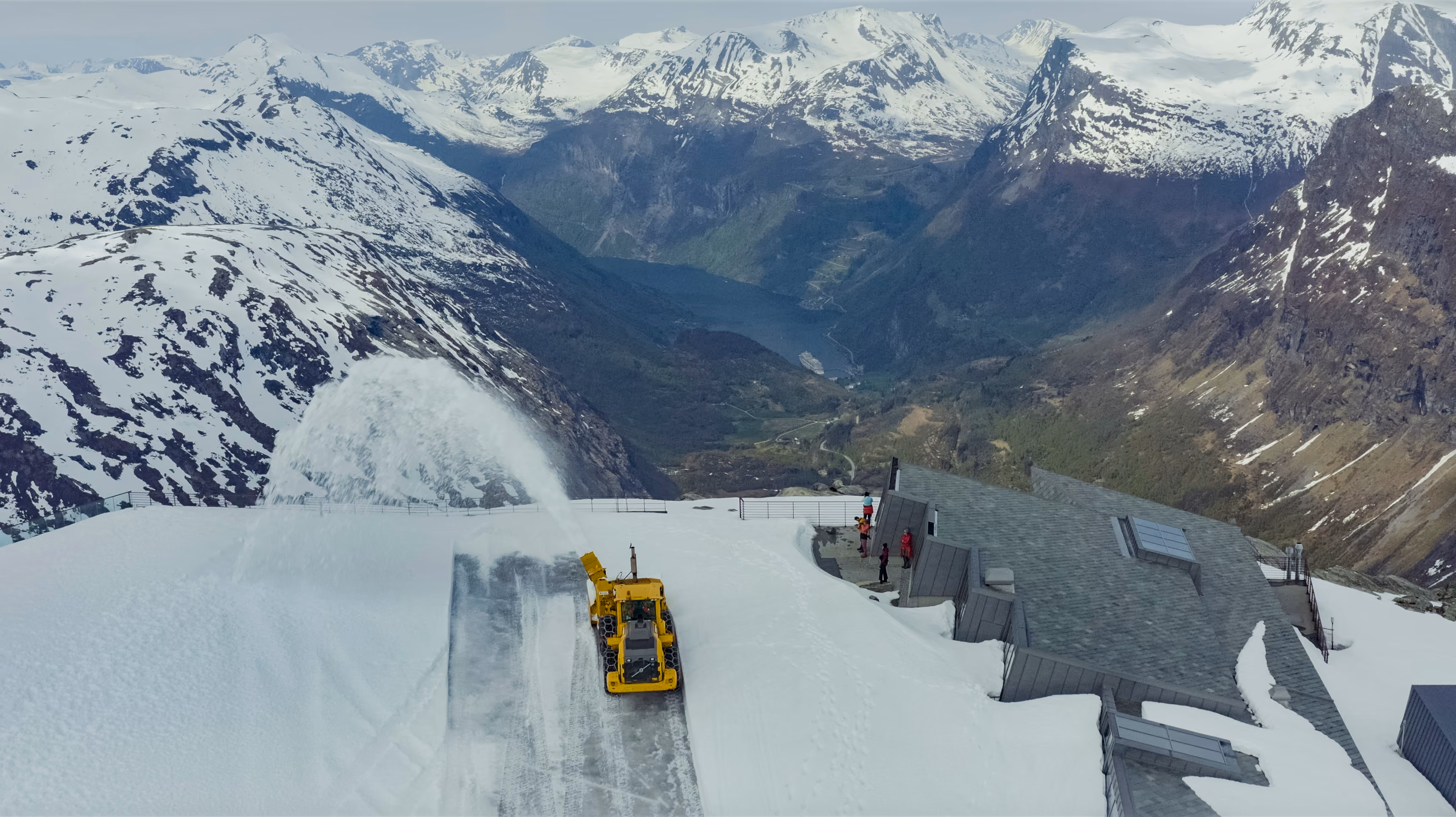 Yellow snowplow clearing snow on a mountain road with snow-covered peaks and a valley in the background.