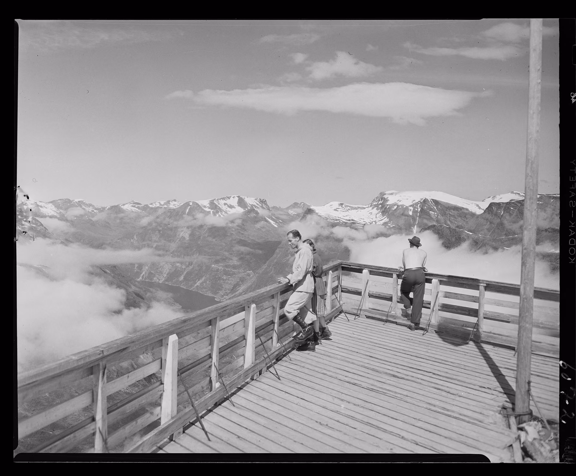 Three people standing on a wooden observation deck overlooking snow-capped mountains and clouds.