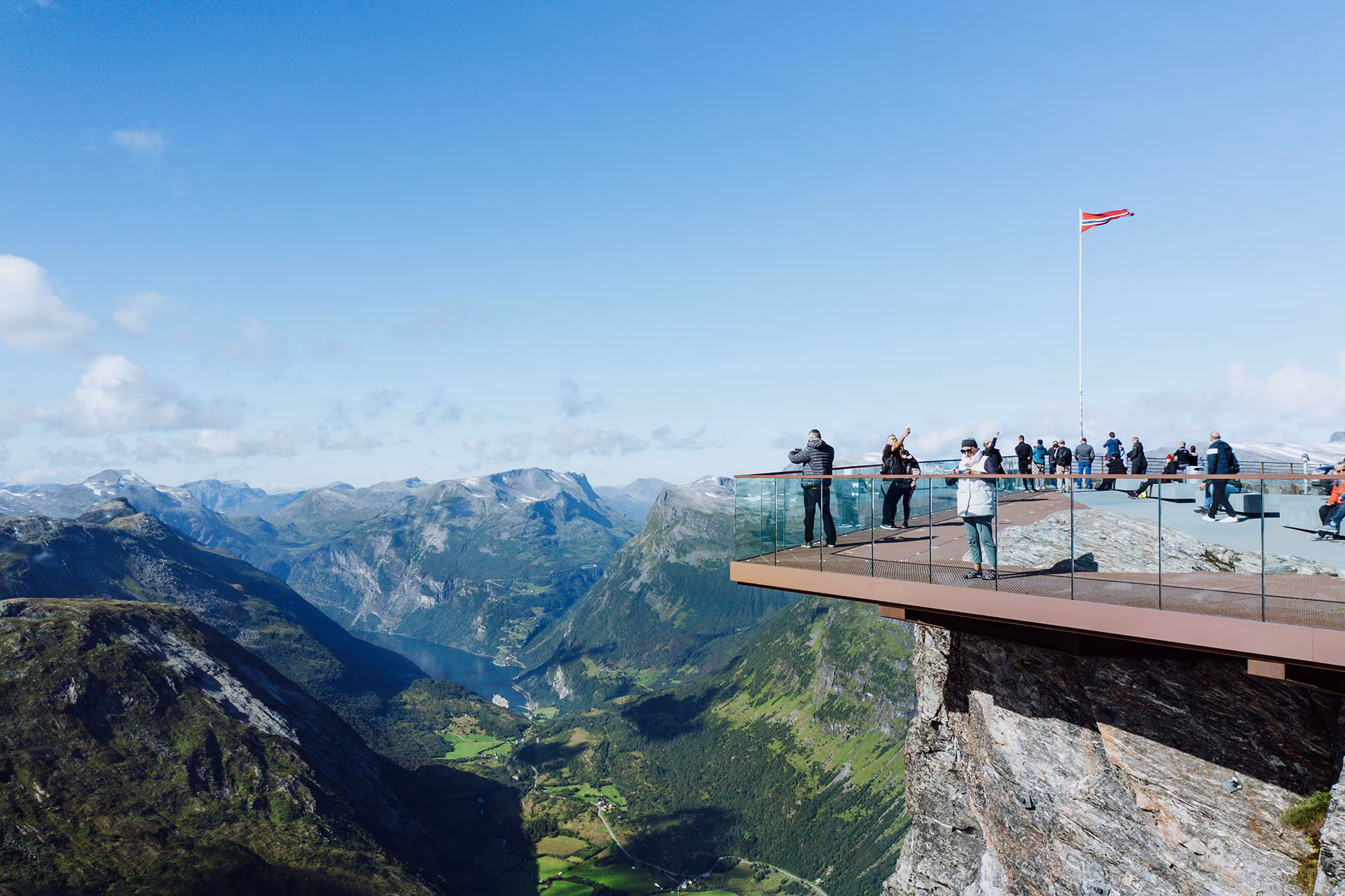 Tourists on a glass viewing platform overlooking a green valley with mountains and a fjord under a clear blue sky.