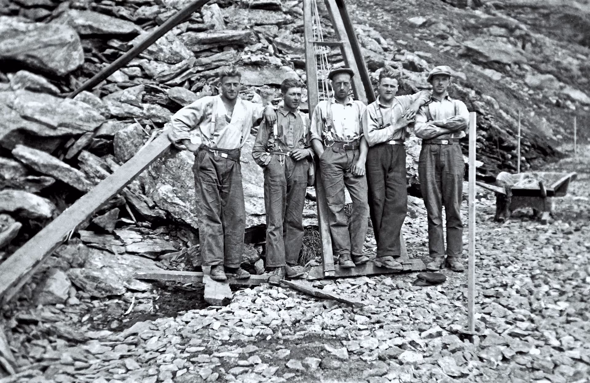 Five men in work clothes standing on rocky terrain, posing beside wooden construction beams.