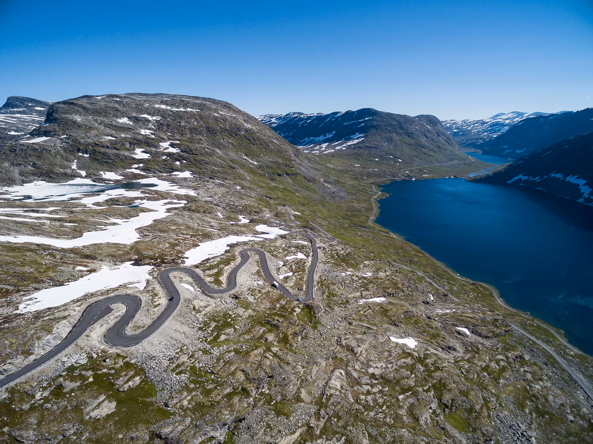 A winding mountain road with several sharp hairpin turns beside a deep blue lake and rocky terrain with patches of snow.