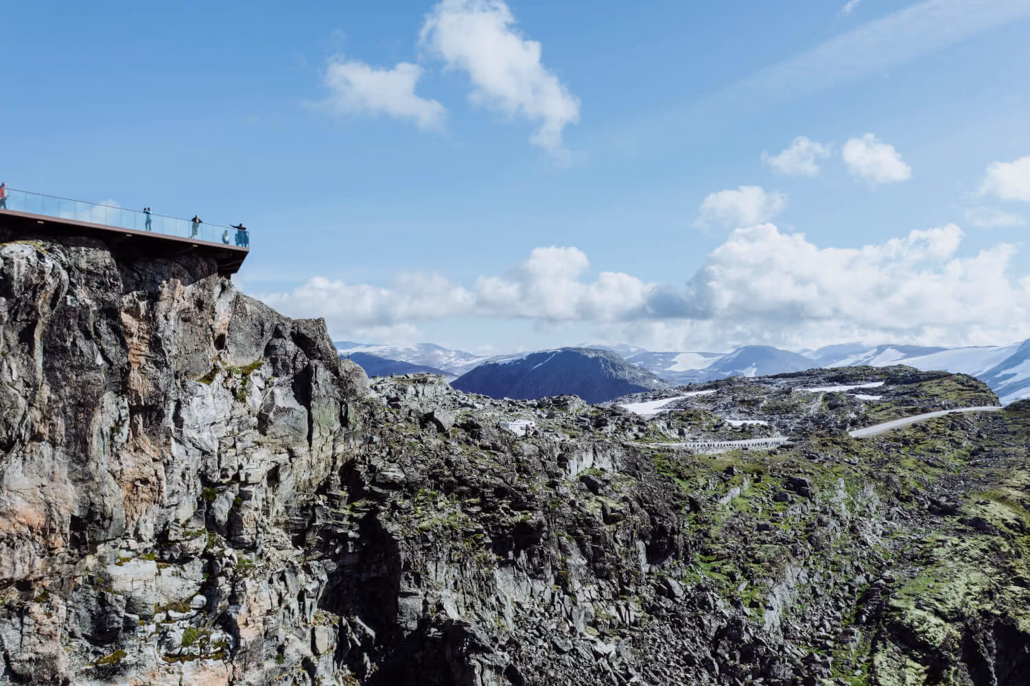 Rocky mountain landscape with a viewing platform on the cliff's edge and snow-capped mountains in the background under a partly cloudy sky.