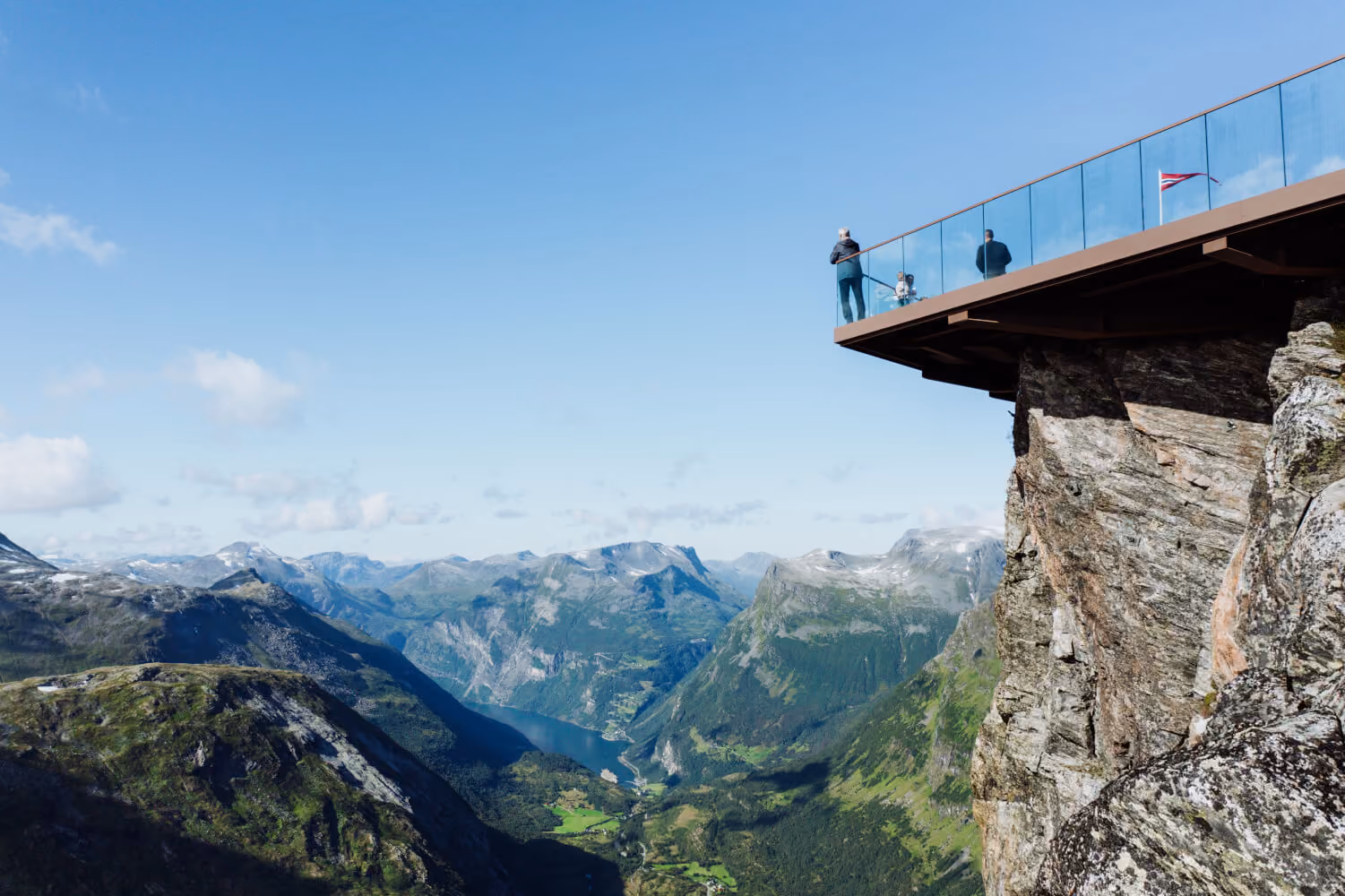 Viewing platform with glass railing and people overlooking a scenic fjord surrounded by mountains under a clear blue sky.