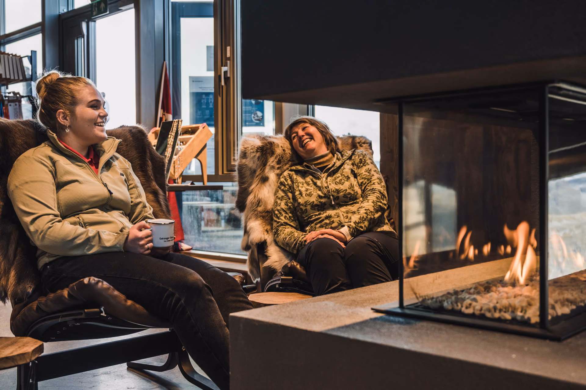 Two women sitting comfortably on fur-covered chairs, smiling and chatting by a modern indoor fireplace.