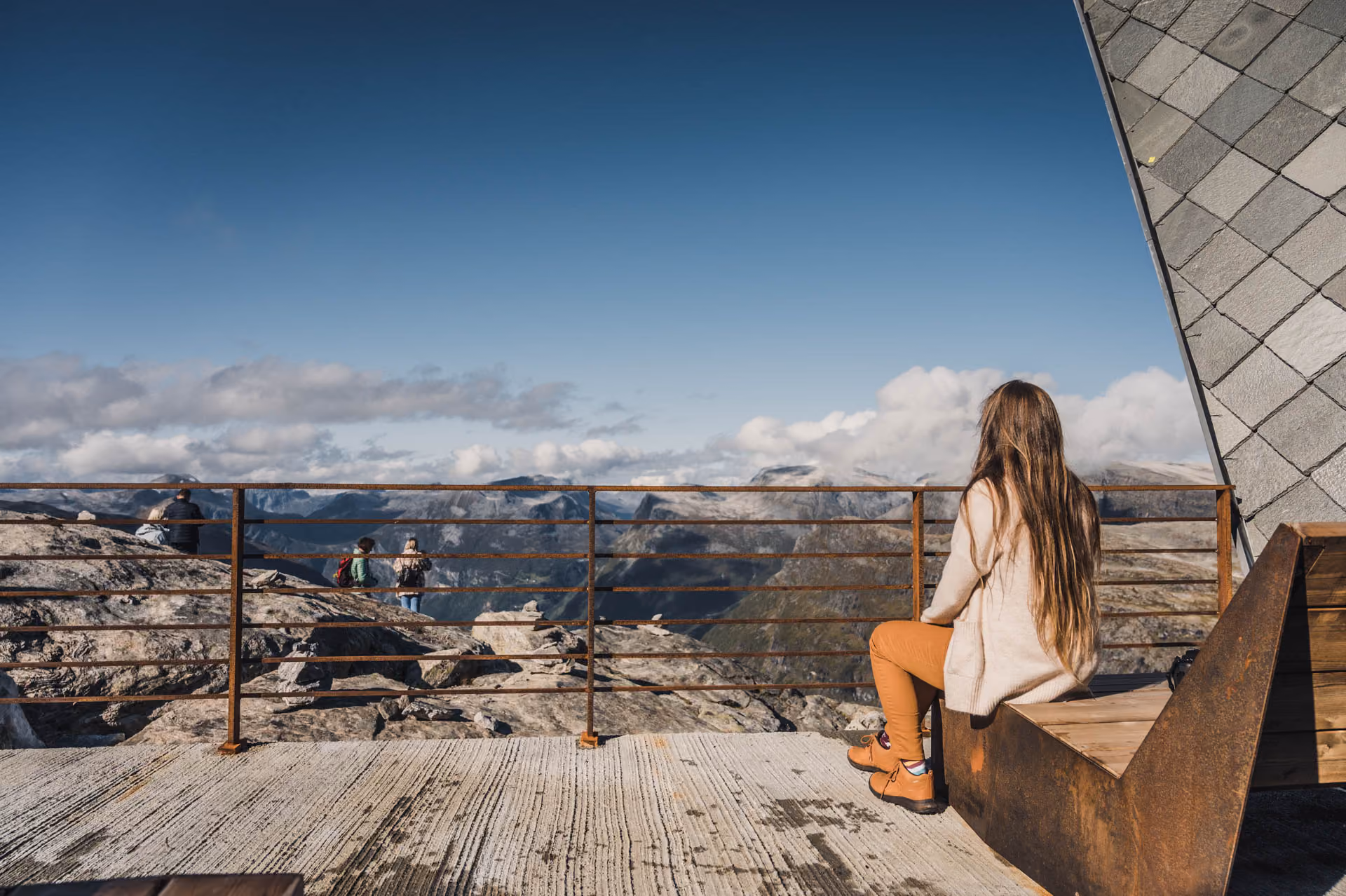 Woman with long hair sitting on a metal and wooden bench, overlooking rocky mountains under a clear blue sky.