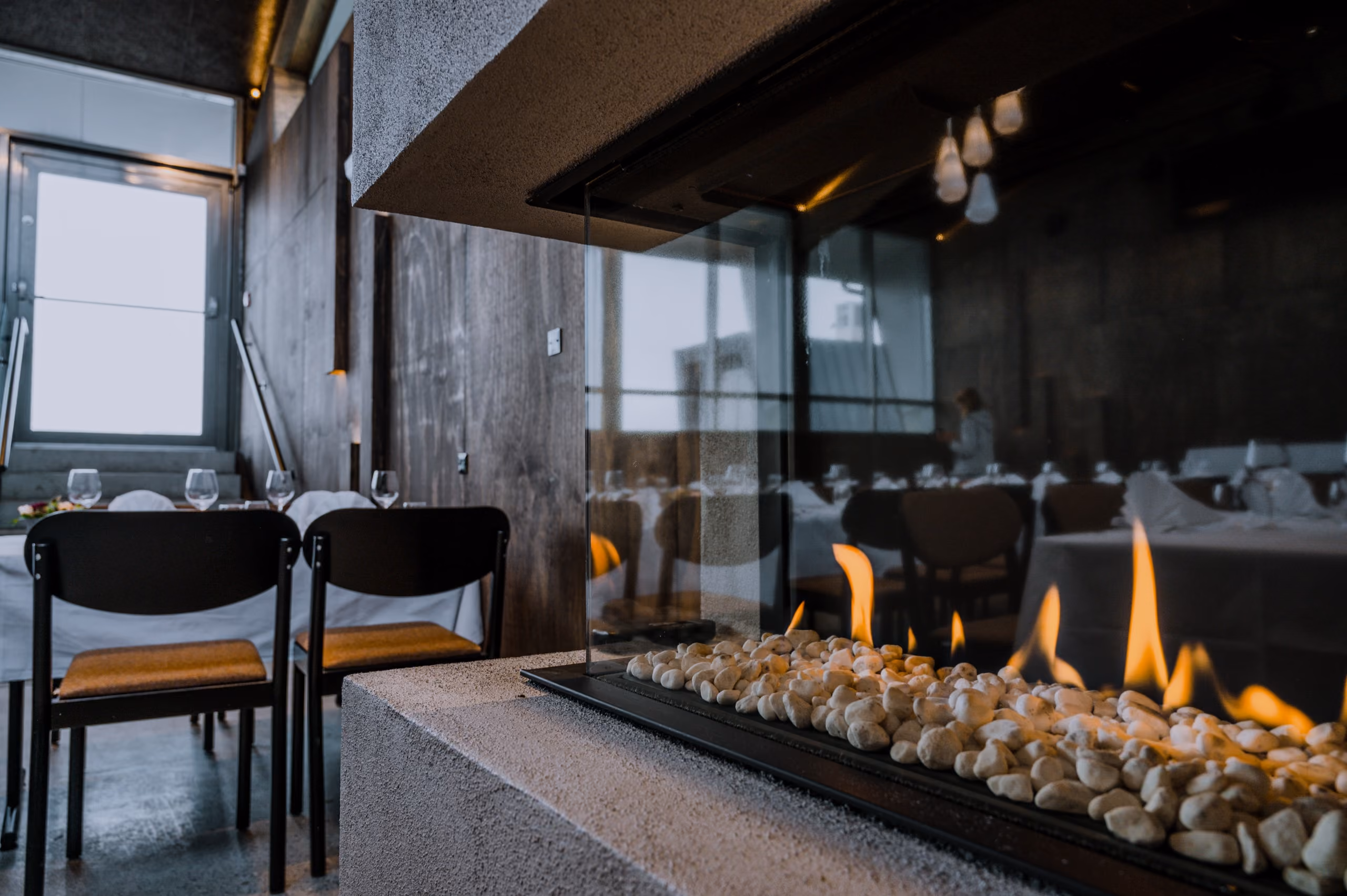 Modern restaurant interior with a glass-enclosed fireplace and dining tables with white tablecloths.
