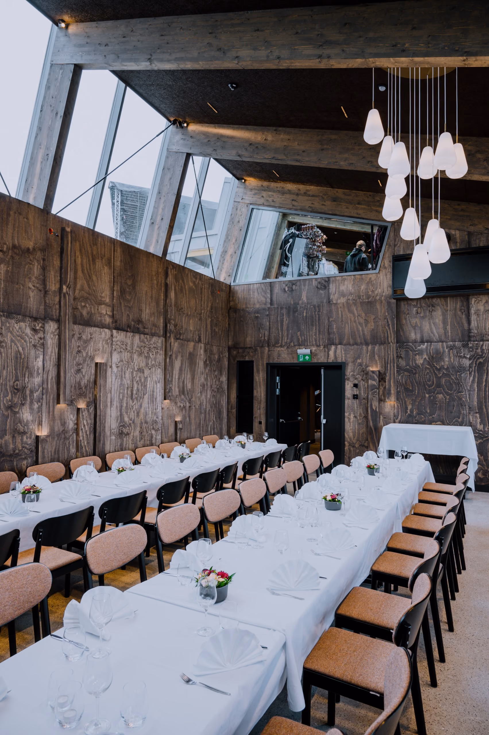 Modern dining room with two long tables, white tablecloths, chairs, folded napkins, and hanging lights.
