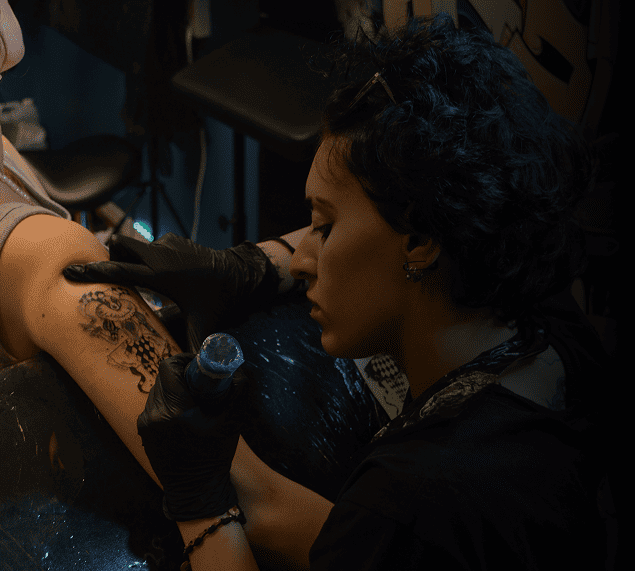 A  tattoo artist  with short dark hair focuses intently while applying a detailed musical note tattoo on a client's arm using a tattoo machine.