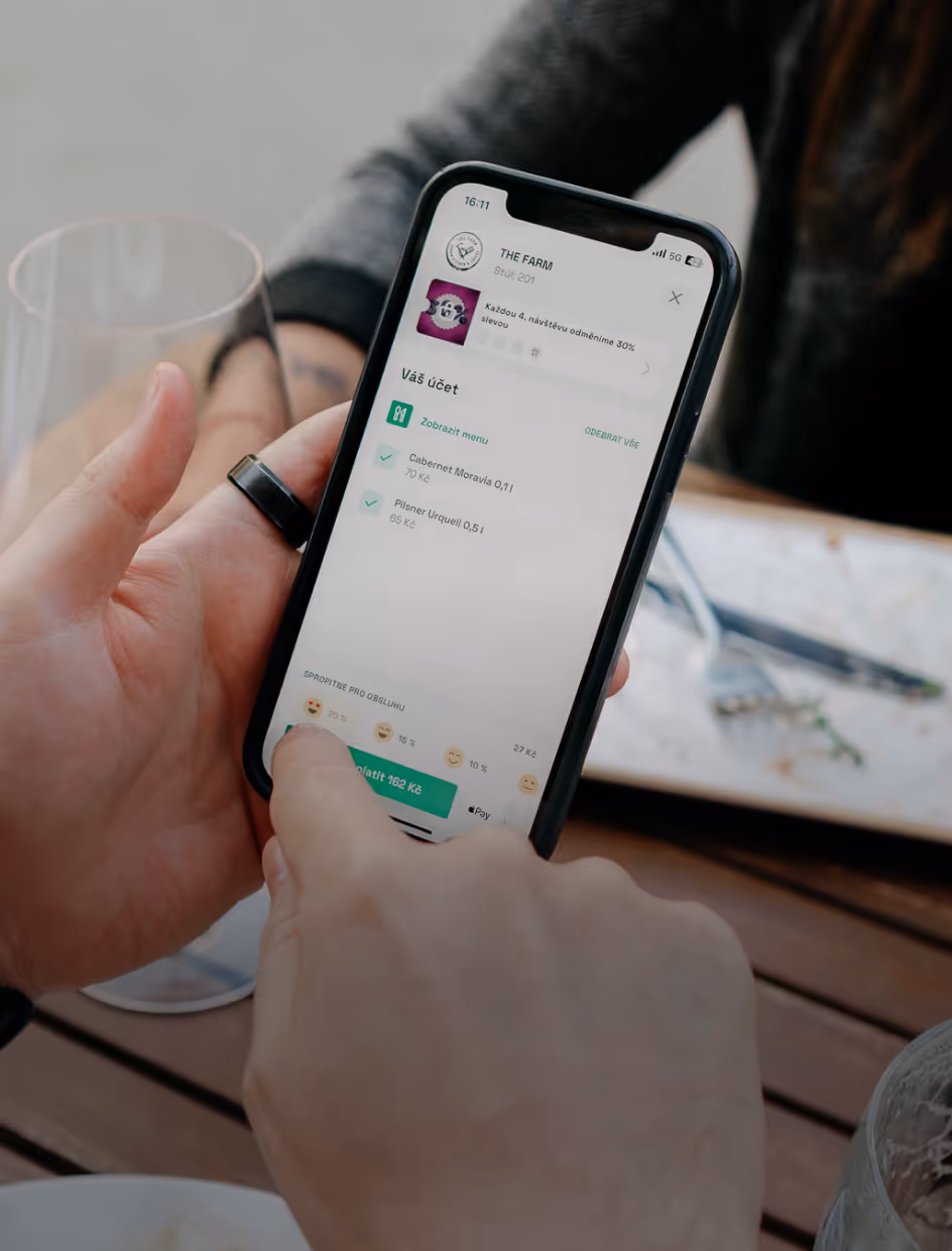 Osoba drží smartphone se zobrazením digitálního účtu v restauraci s objednávkou dvou nápojů a možností přidání spropitného.