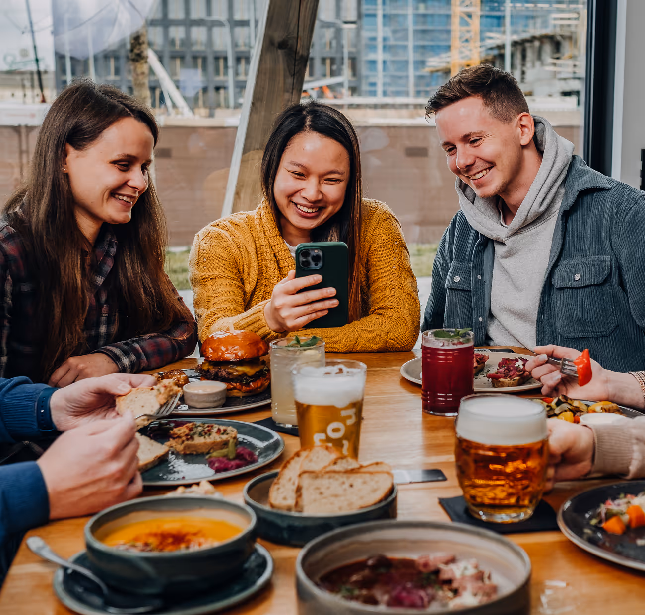 Skupina usměvavých přátel sdílí jídlo a pití u stolku v restauraci, zatímco jedna žena ukazuje něco na mobilním telefonu.