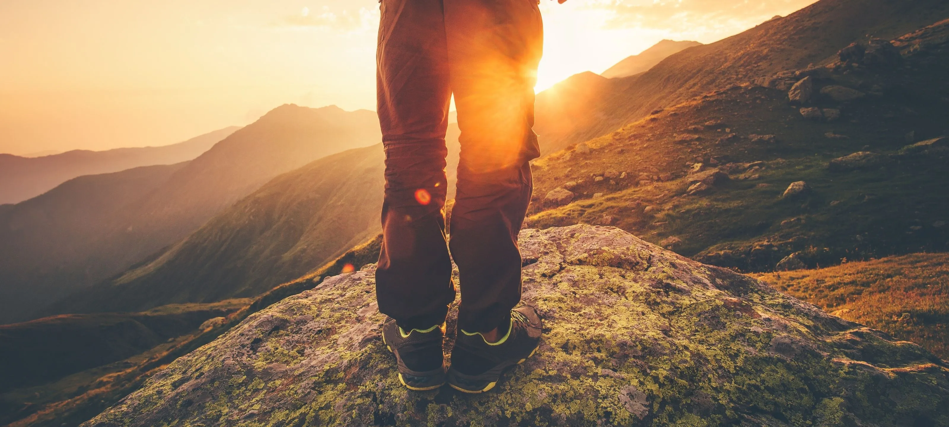 Mann beim Wandern, steht auf einem Felsen während des Sonnenuntergangs – nur die Beine sichtbar‍