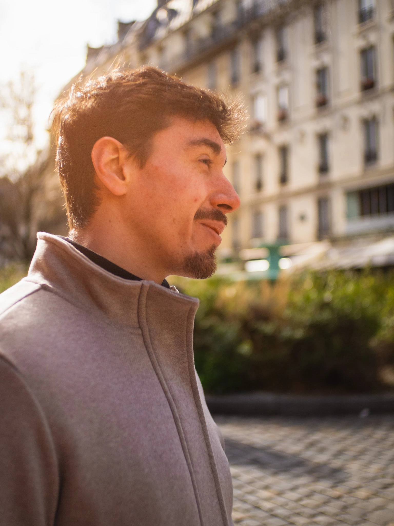 Man with short dark hair and goatee wearing a light brown jacket outdoors with buildings and greenery in the background.