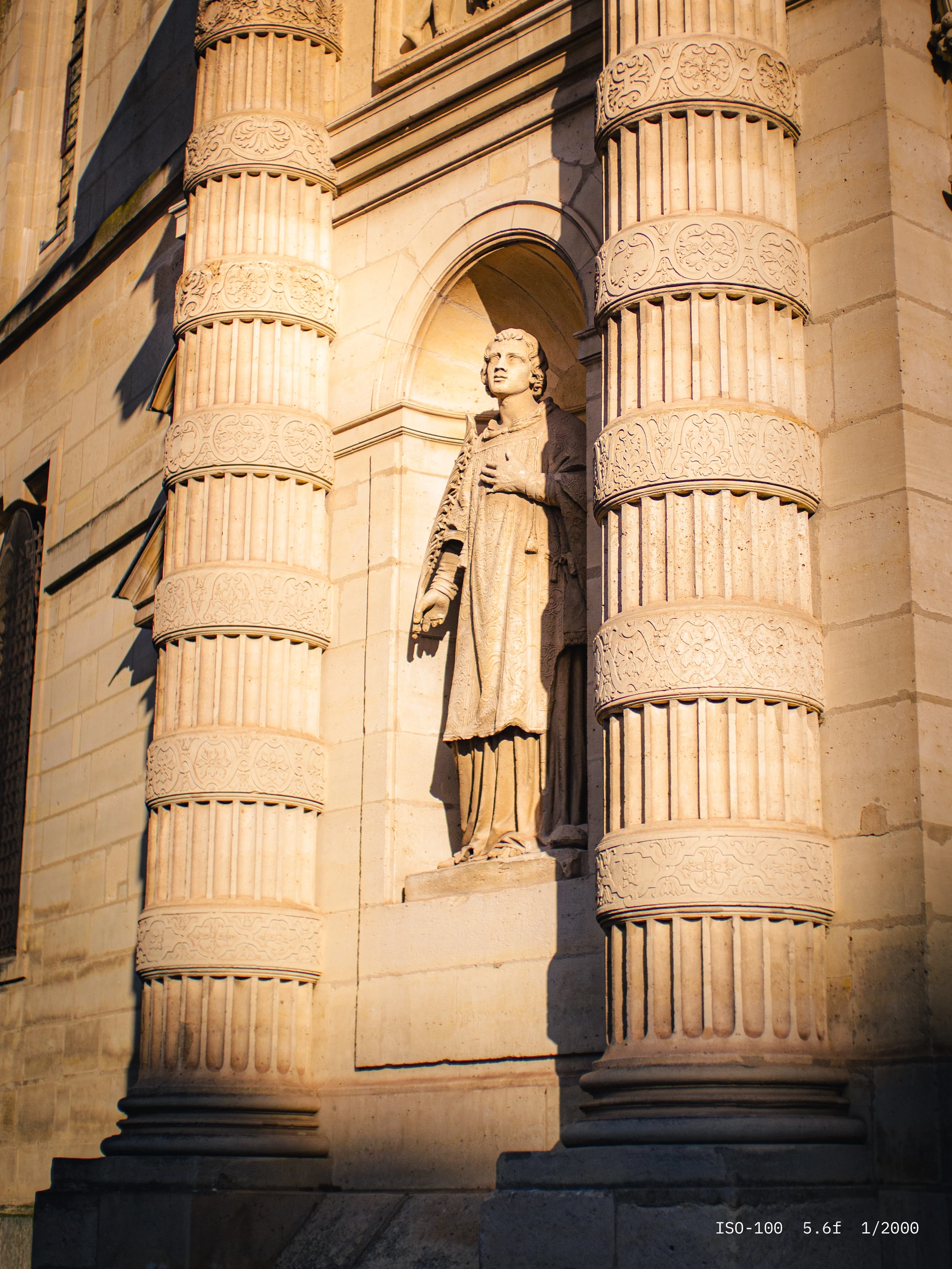 Stone statue of a robed figure standing between two ornately carved columns on a building facade.