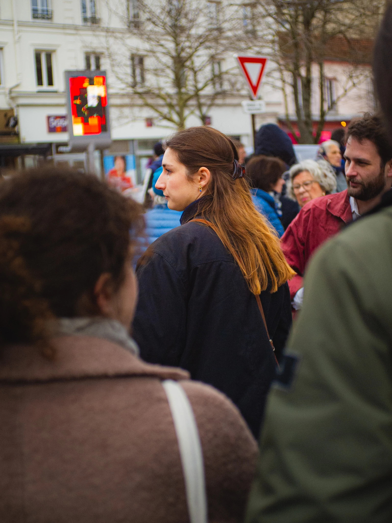 Crowd of people walking down a city street in cool weather, focusing on a young woman with long brown hair looking to the left.