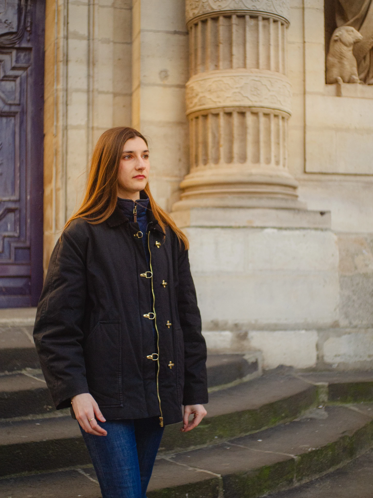 Young woman with long brown hair wearing a black coat and blue jeans standing on stone steps near a carved stone column.
