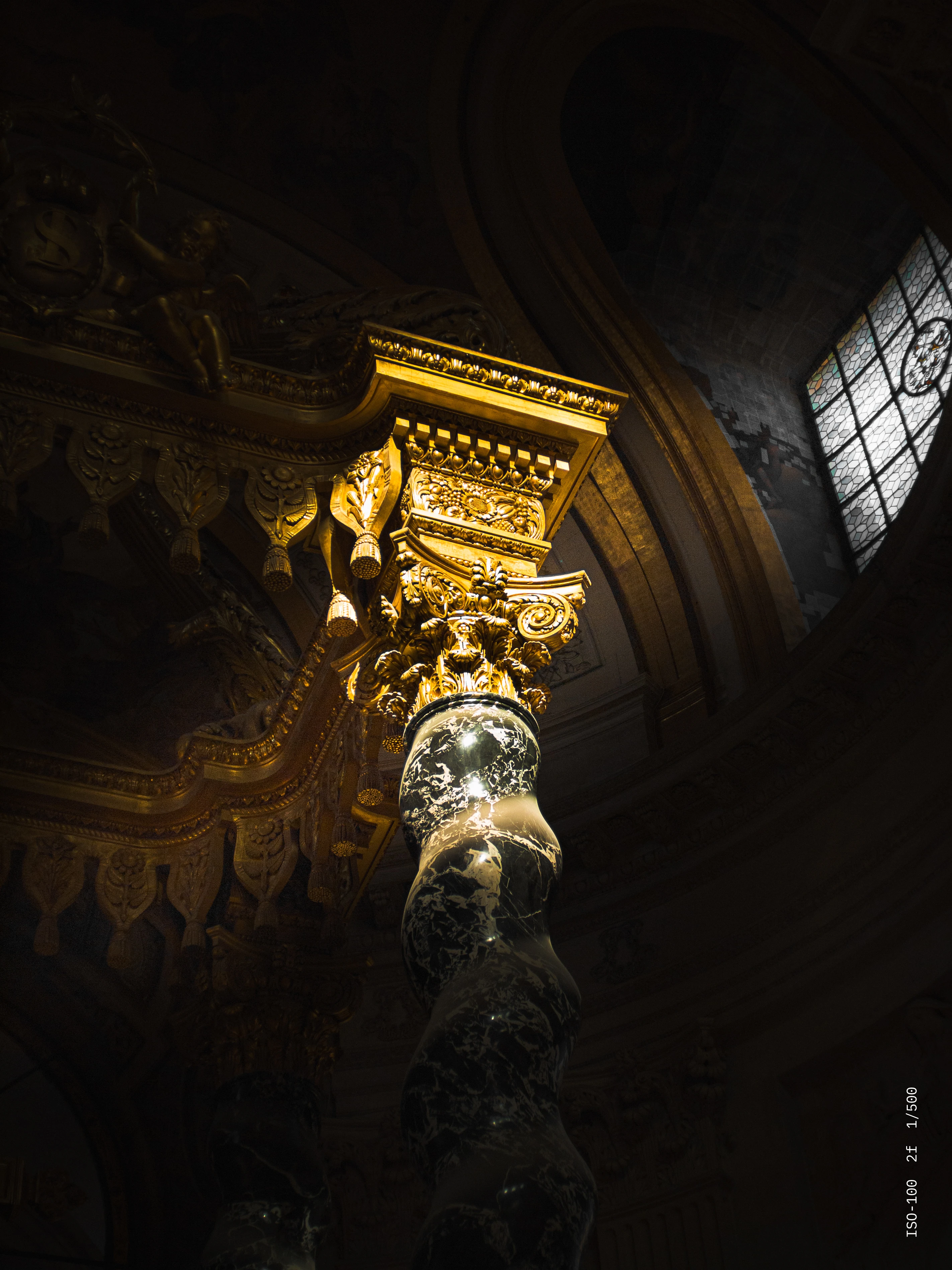 Ornate marble column with gold leaf capital illuminated inside a building with stained glass window.