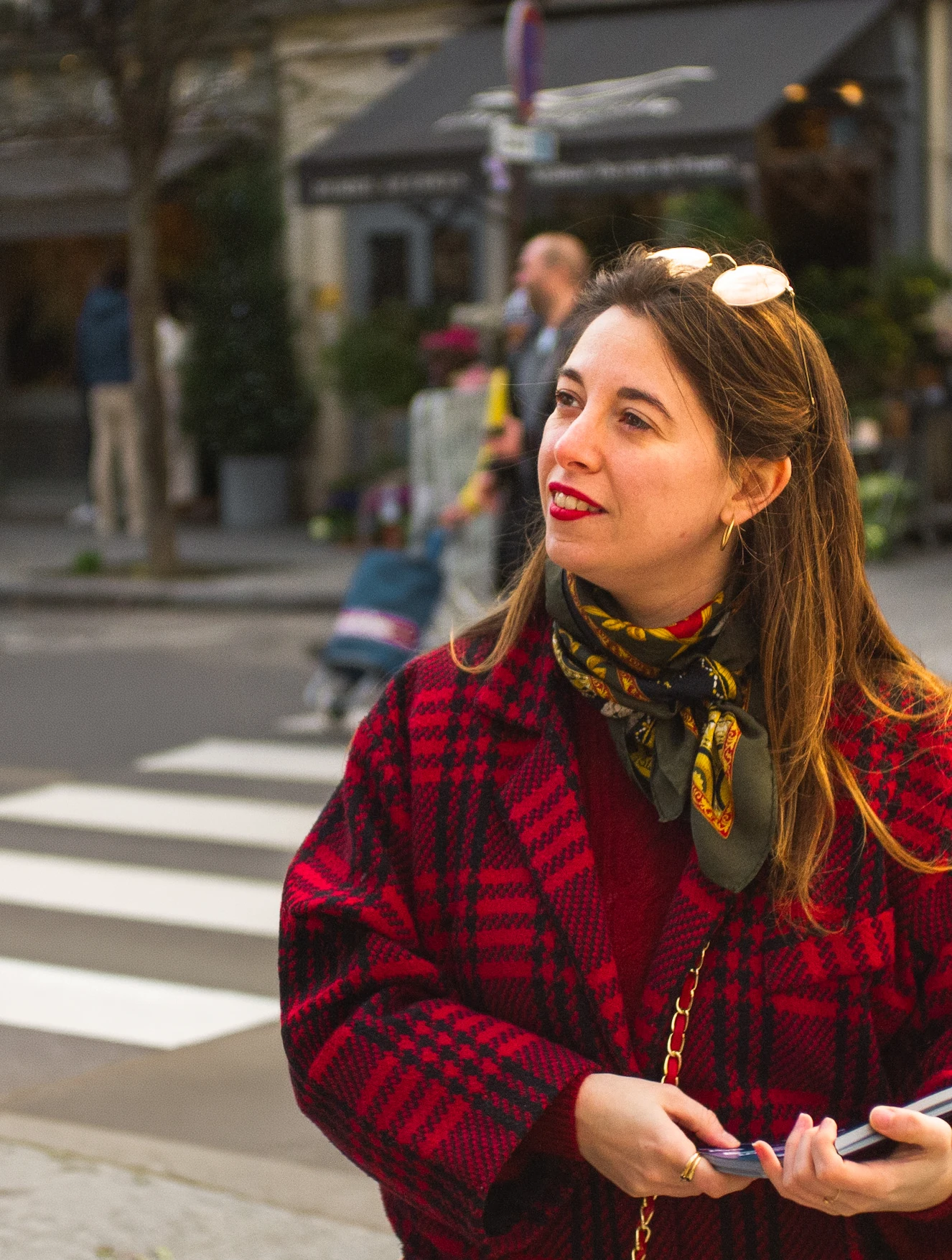 Woman with long brown hair and red lipstick wearing a red plaid coat and patterned scarf, standing near a pedestrian crossing in an urban setting.