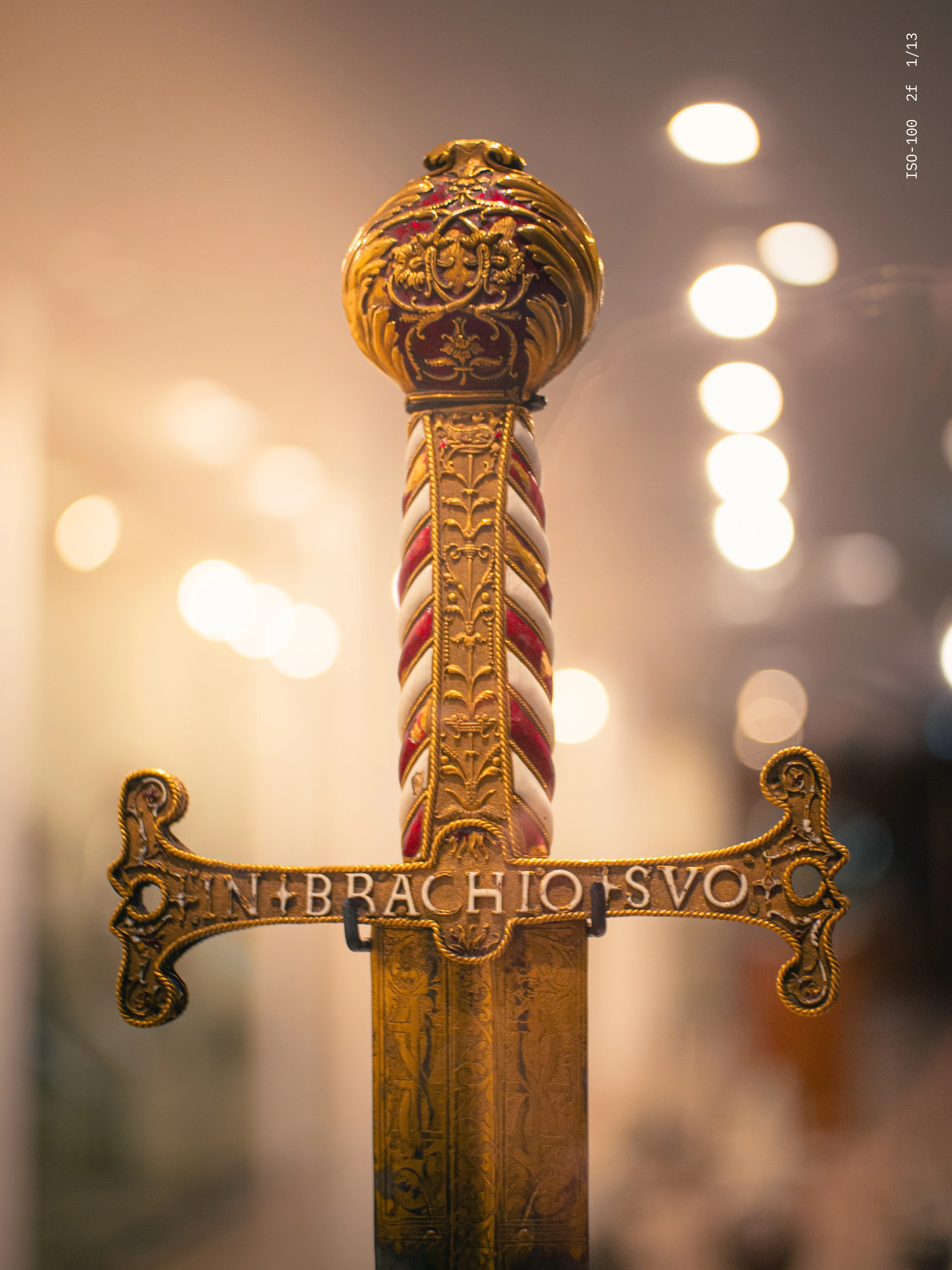 Close-up of an ornate sword hilt with red and white twisted handle and gold detailing, inscribed with 'IN BRACHIO SVO'.