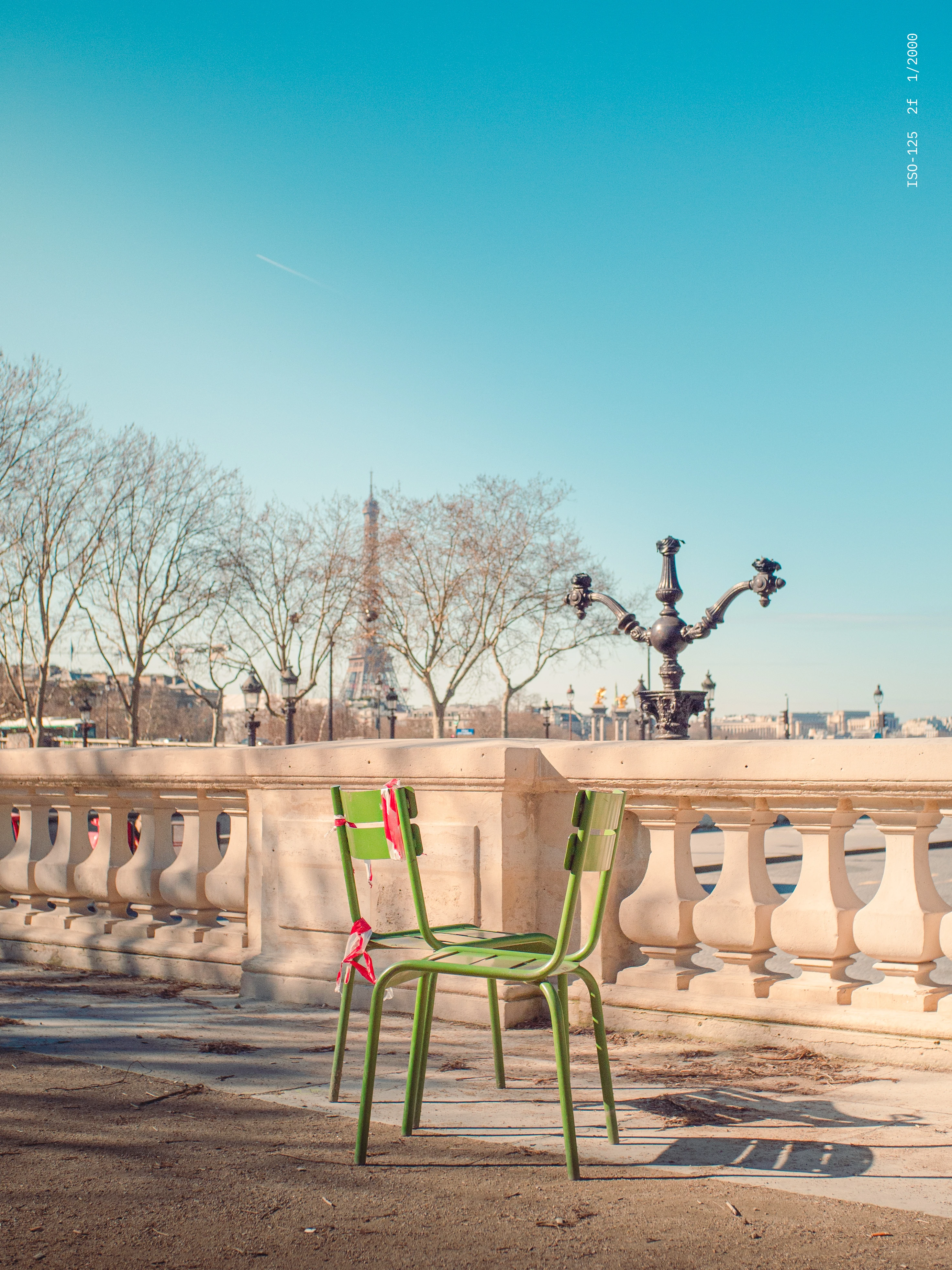 Green metal chair tied with red and white ribbons in front of Parisian balustrade with Eiffel Tower and leafless trees in the background under clear blue sky.
