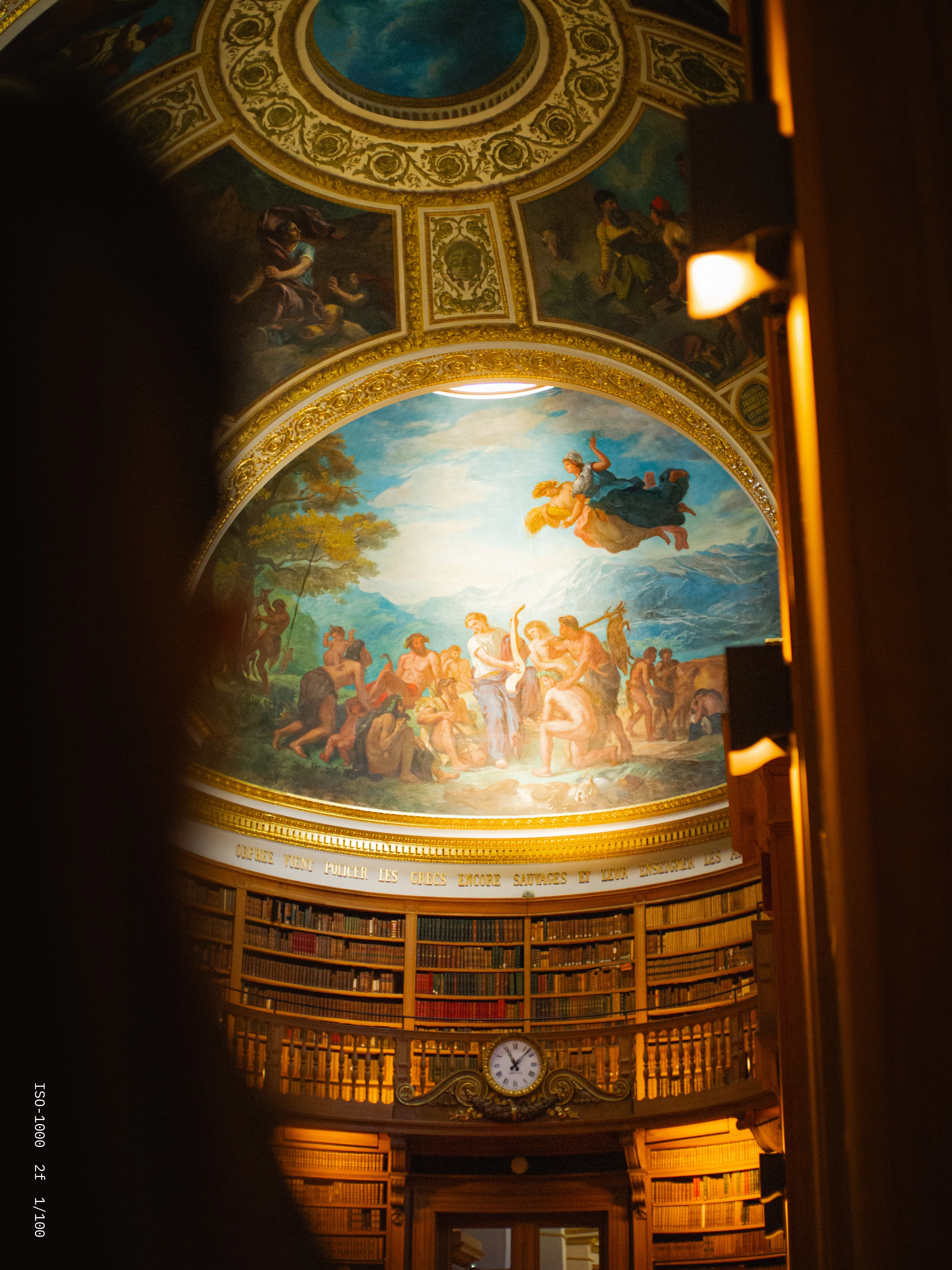 Ornate library interior featuring a large circular ceiling mural with classical figures over wooden shelves filled with books and a clock above an entrance.