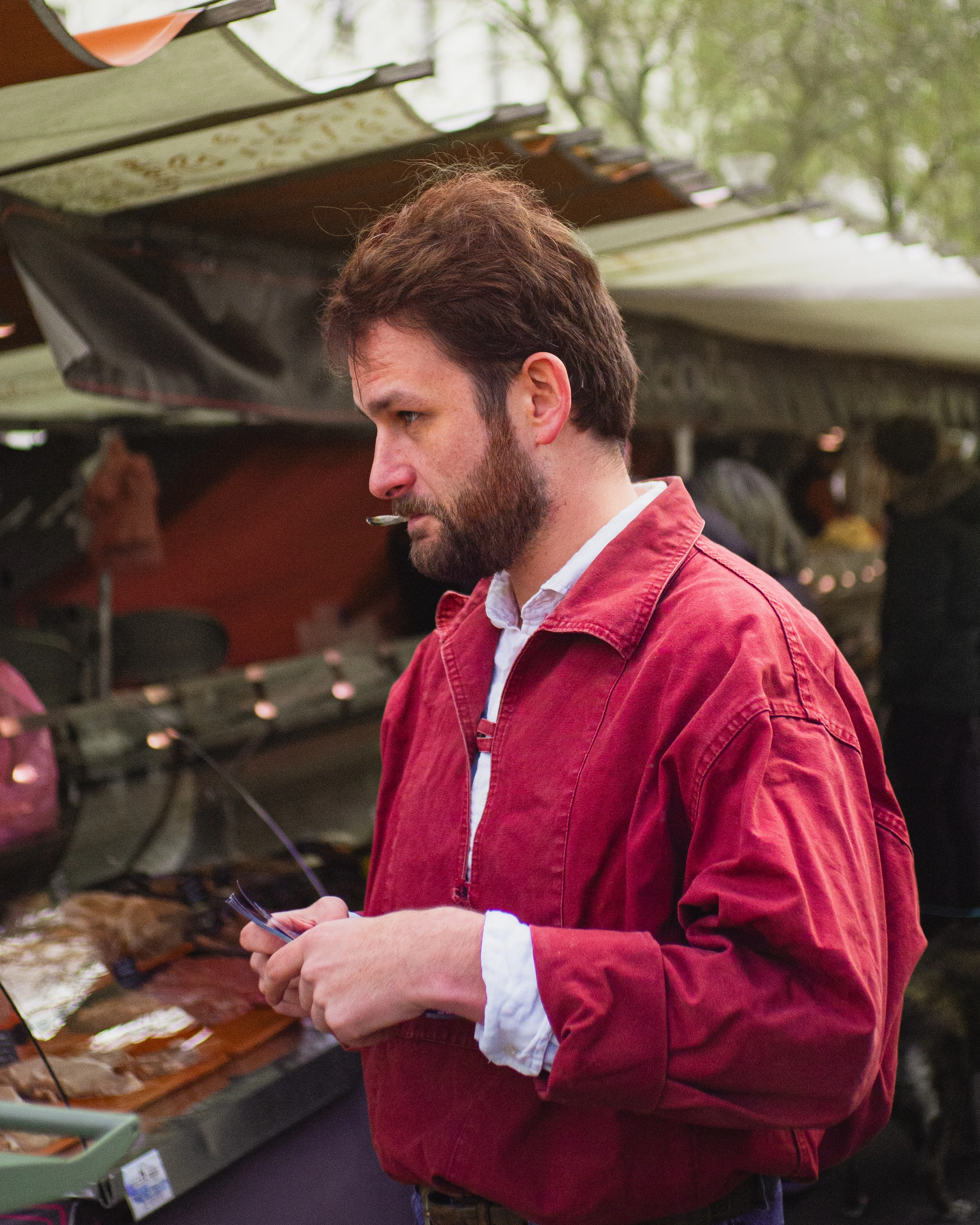 Bearded man in a red jacket at an outdoor market stall.