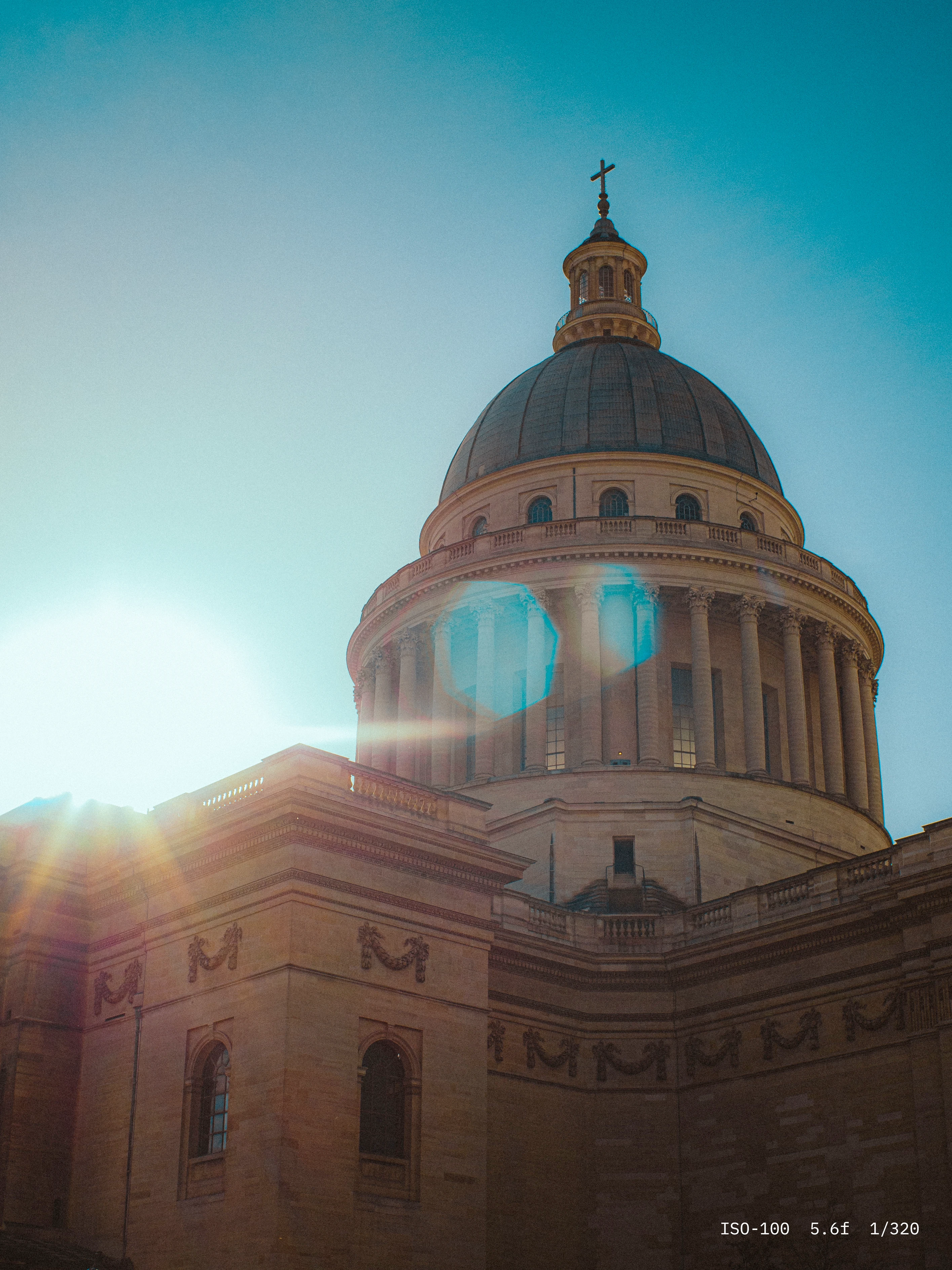Sunlight shining next to a large classical stone dome with columns and a cross on top against a clear blue sky.