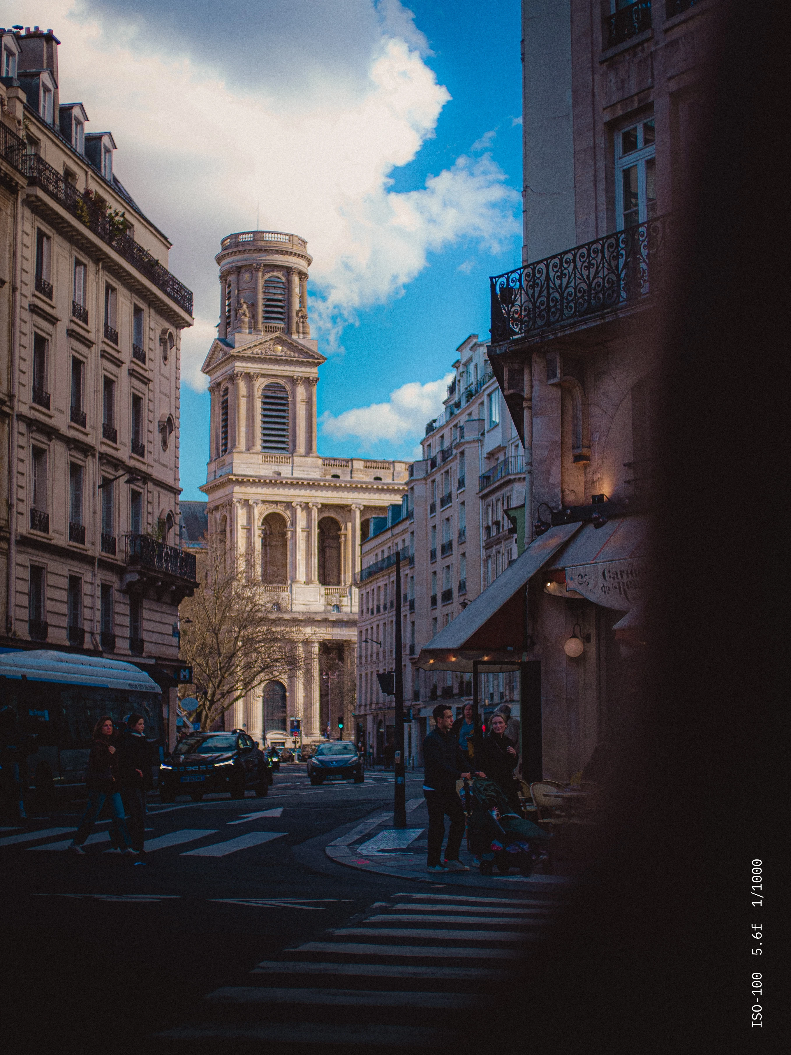 Street view of historic European buildings with a prominent stone tower under a blue sky with clouds.