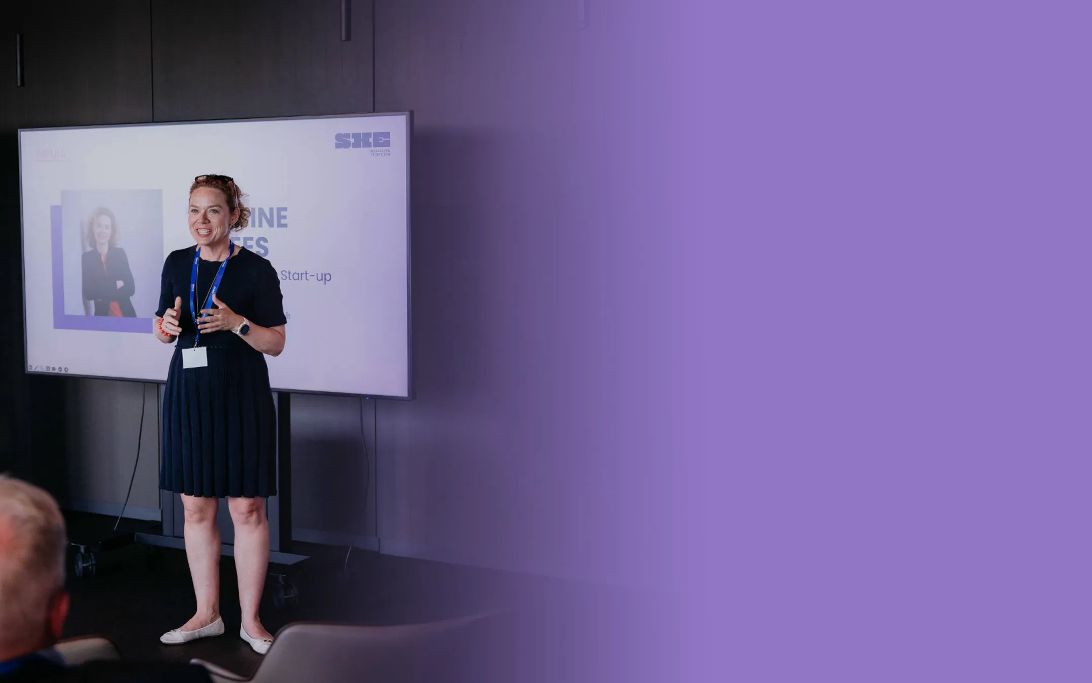 Lächelnde Frau mit braunen Haaren, die einen grauen Blazer trägt, vor einem Hintergrund mit blauen und rosa Formen.