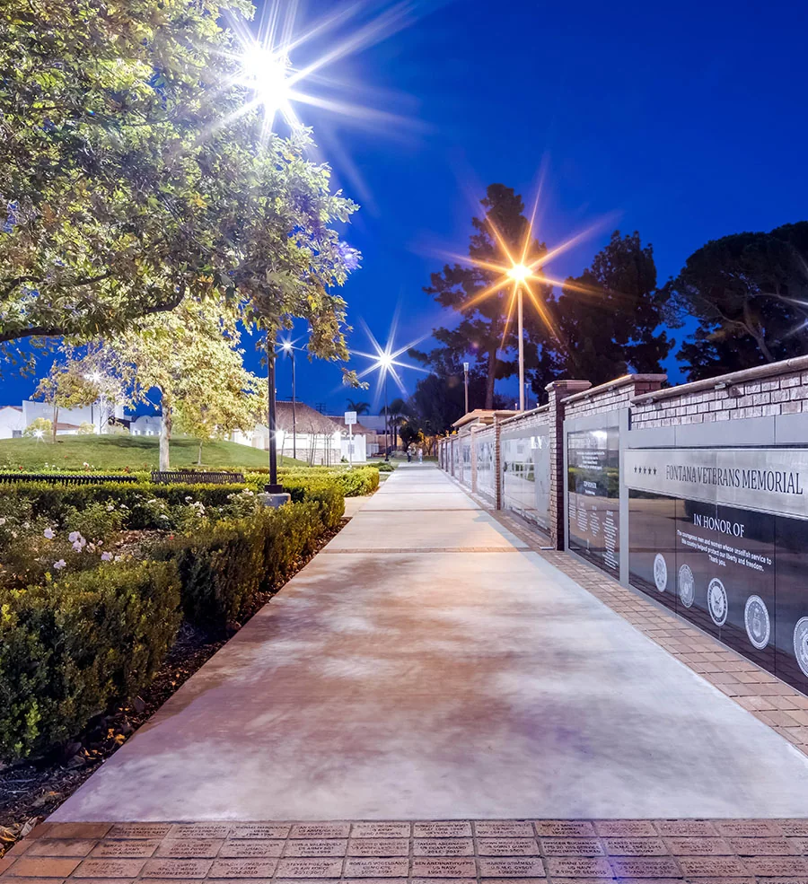 Public works projects: illuminated memorial walkway beside planted beds and a brick wall at night