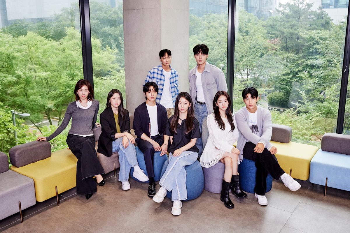 A group of eight young adults, four men and four women posing together indoors in a modern, well lit room with large windows revealing a lush green landscape outside.