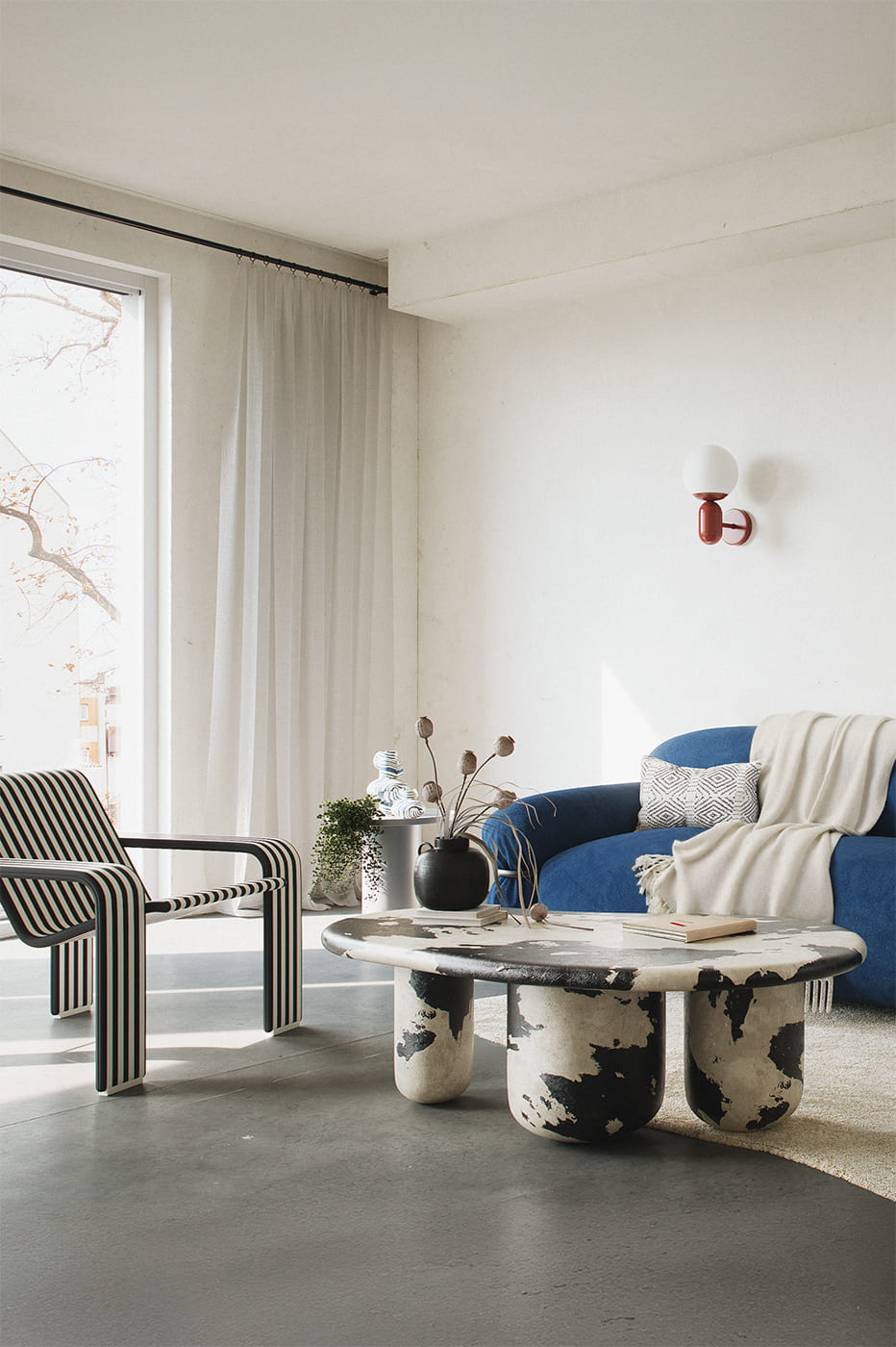 Modern minimalist living room featuring a sculptural black and white coffee table, a bold blue curved sofa with a neutral throw blanket, and a striped designer armchair. Natural light fills the space through large windows with sheer curtains. Decorative vases and dried flowers add a touch of organic texture.