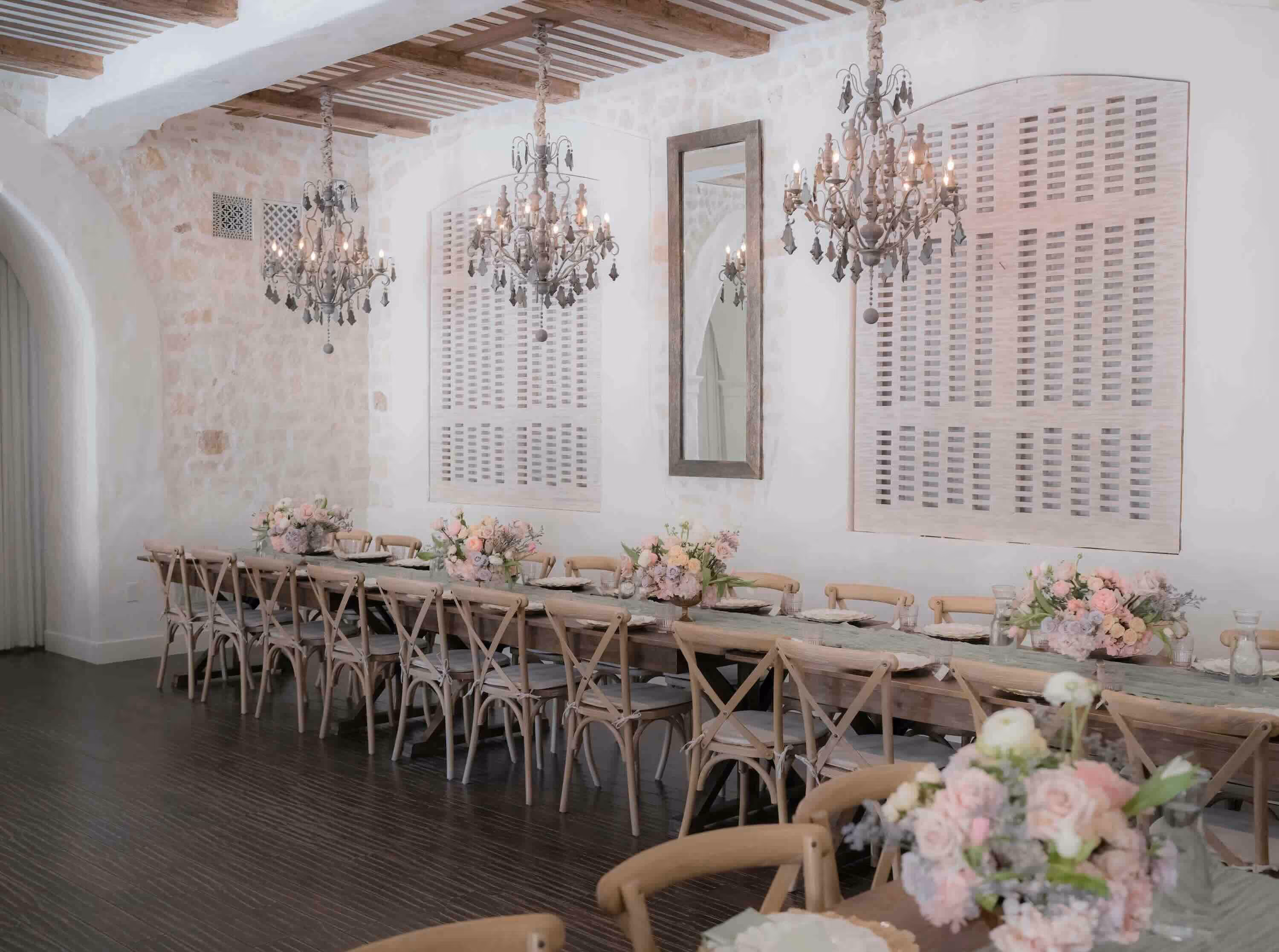 Long dining tables in Santuari LA Main Room decorated with soft pink and white floral centerpieces, chandeliers, and stone walls for luxury weddings and private events in Toluca Lake