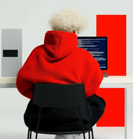 Person with curly blonde hair wearing a red hoodie sitting at a desk working on a computer with code on the screen.