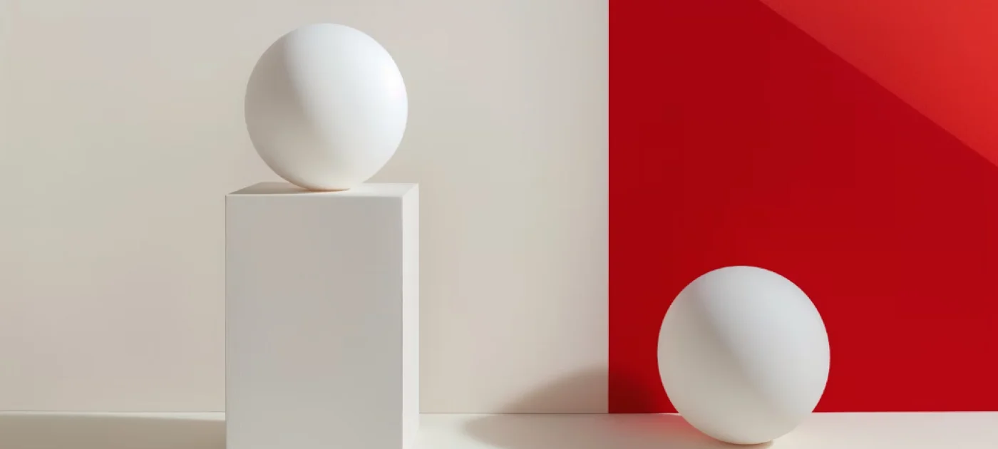 Two white spheres casting shadows, one on a tall white rectangular pedestal against a beige wall, and another resting on a white surface near a red wall corner.
