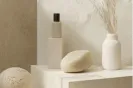 Minimalist beige-toned decor featuring a round textured object, a white bottle, a beige stone, and a white vase with dried pampas grass on a white shelf.