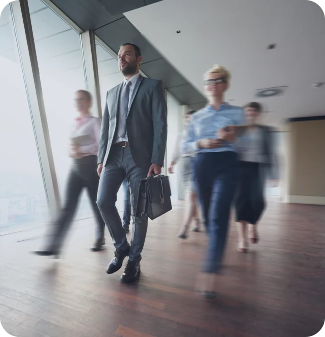 Businessman in grey suit carrying leather briefcase walking purposefully through modern office with floor-to-ceiling windows. Other professionals appear in motion blur behind him, creating sense of dynamic business environment