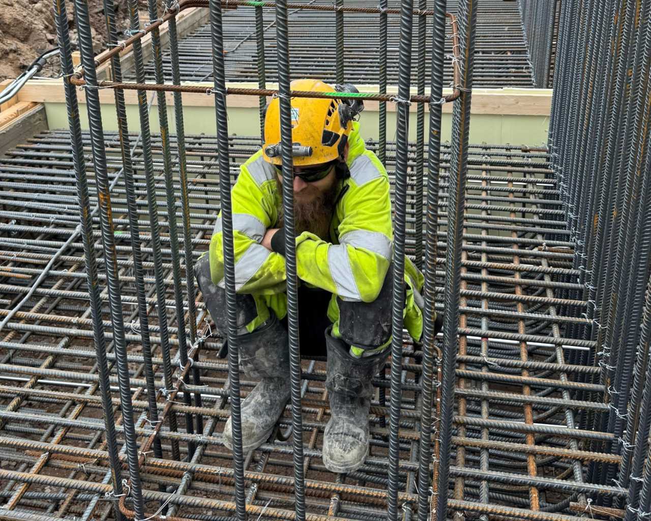 Rakennustyöntekijä keltaisessa suojakypärässä ja heijastinvaatteissa istuu betoniteräksistä rakennetussa kehässä.