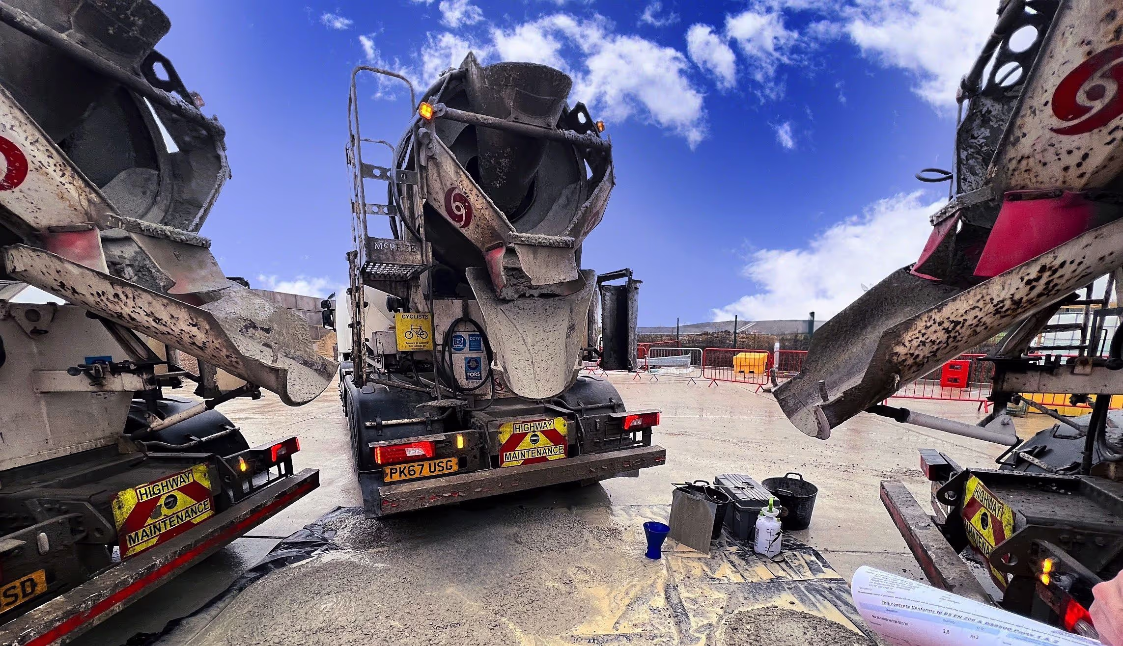Three concrete mixer trucks with chutes extended parked on a construction site under a blue sky with some clouds.