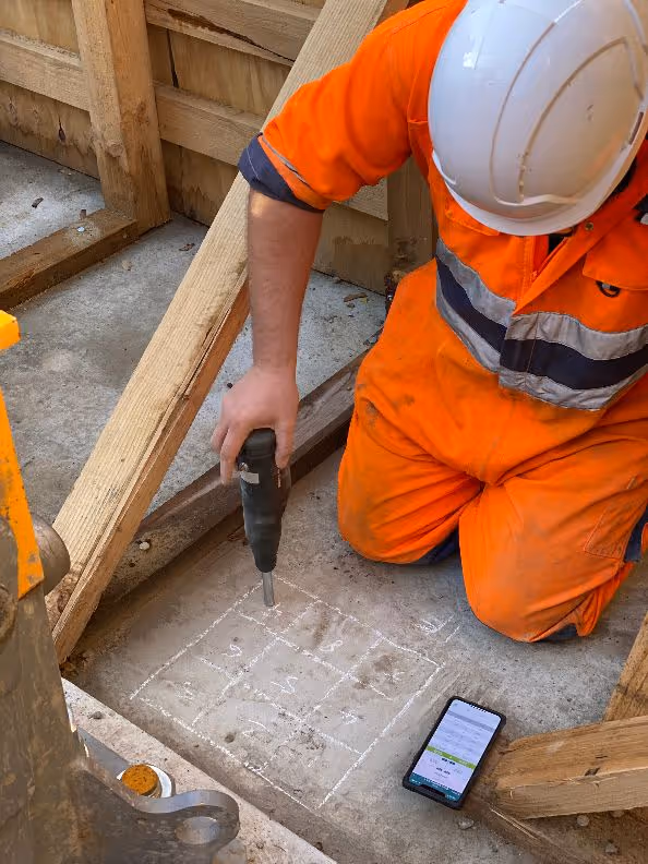A technician in orange safety gear and a white helmet kneels on a concrete floor. They are testing the strength with a Schmidt rebound hammer.