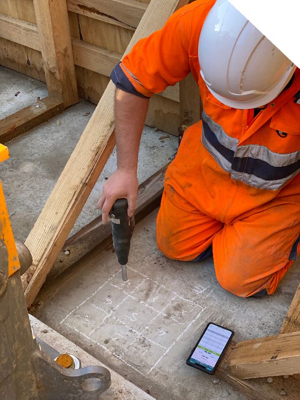 A technician in orange safety gear and a white helmet kneels on a concrete floor. They are testing the strength with a Schmidt rebound hammer.